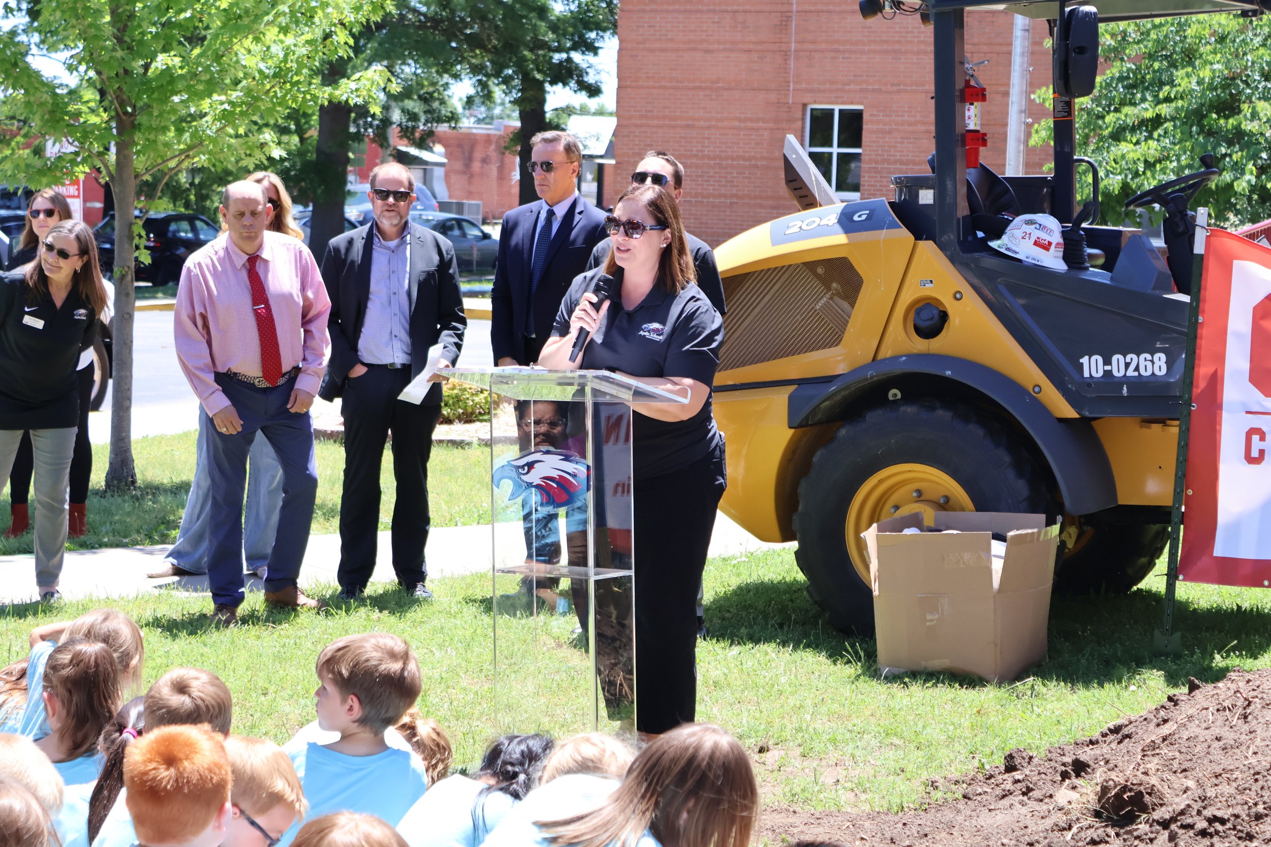 A woman speaking at a podium during an outdoor event, with a group of people and a yellow construction vehicle in the background, while children sit on the grass nearby.