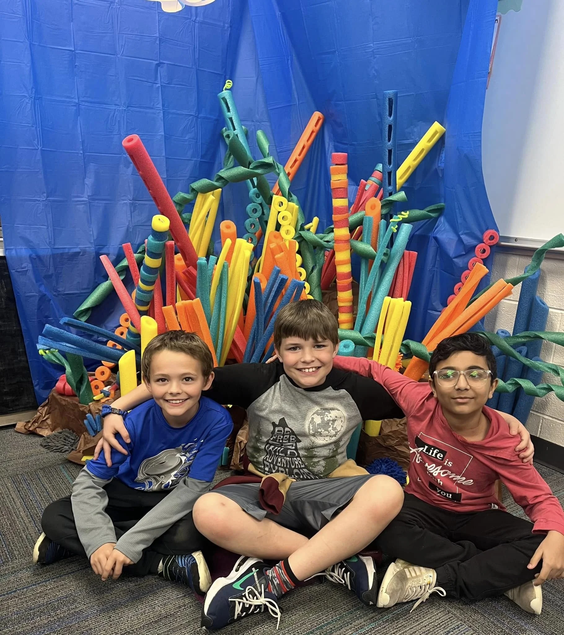 Three boys smiling and sitting in front of a colorful paper craft display of large foam noodles and shapes, with a blue backdrop and white walls.