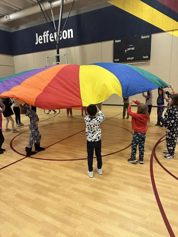 Children in a gymnasium playing with a large, multicolored parachute. The parachute is blue, green, yellow, orange, and red. Some children are holding the edge, lifting it, while others are underneath or around it.