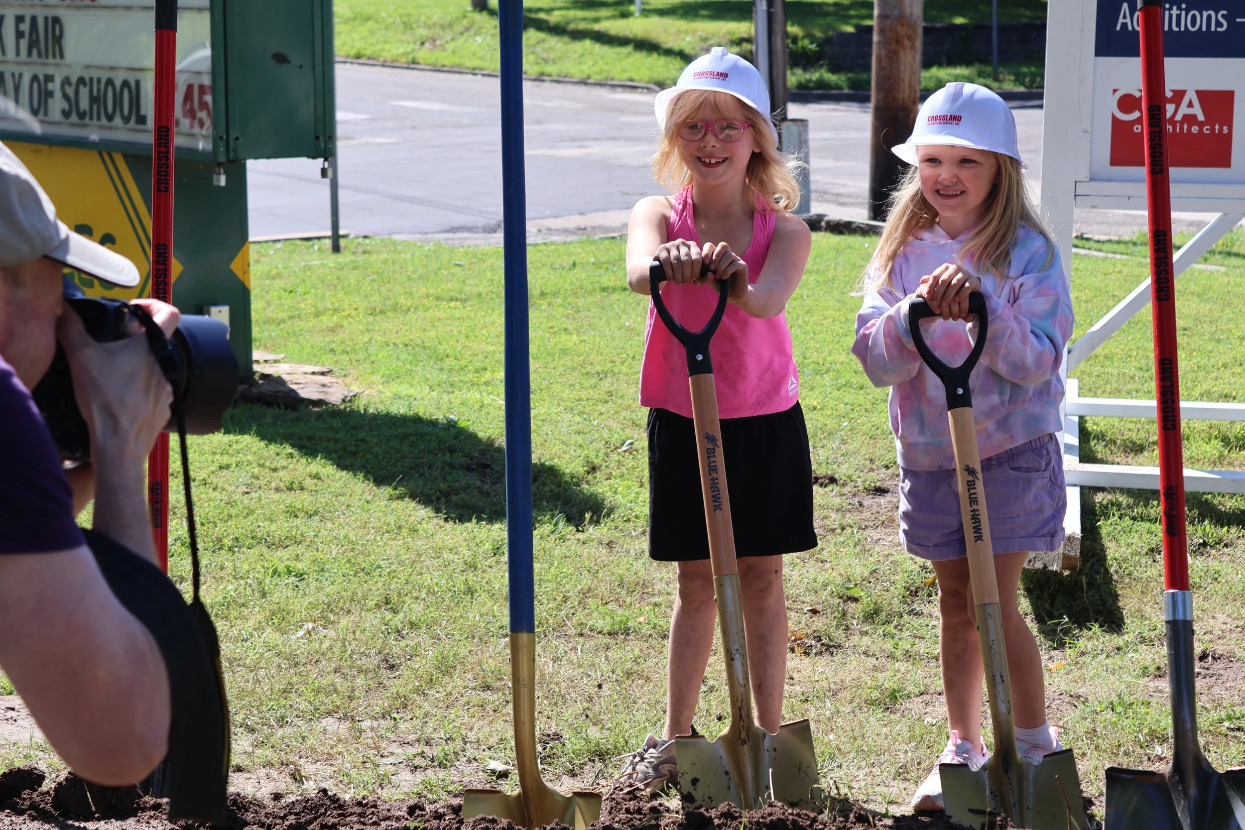 Two young girls with blonde hair, wearing white hats and colorful clothing, holding shovels during a ground-breaking ceremony at a community event.