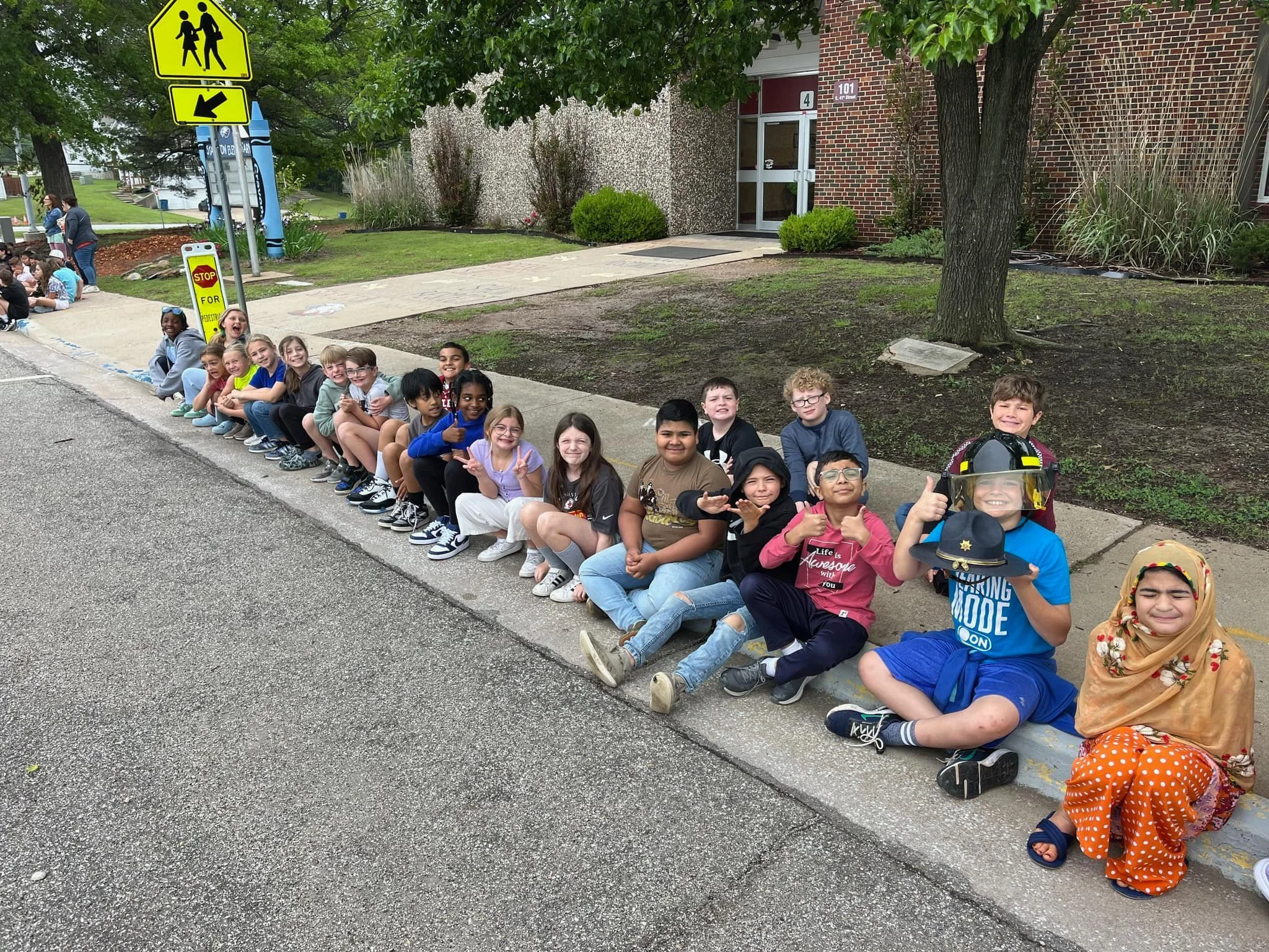 A group of children sitting on a sidewalk outside a school building, some dressed in costumes, smiling and posing for the photo.