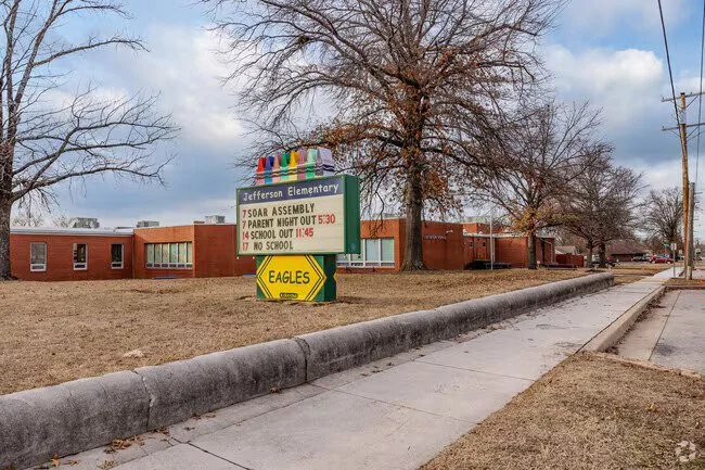 The exterior of Jefferson Elementary School with a colorful rainbow banner, trees, and a lawn. There is a sign showing school events and a yellow arrow-shaped sign for the Eagles.