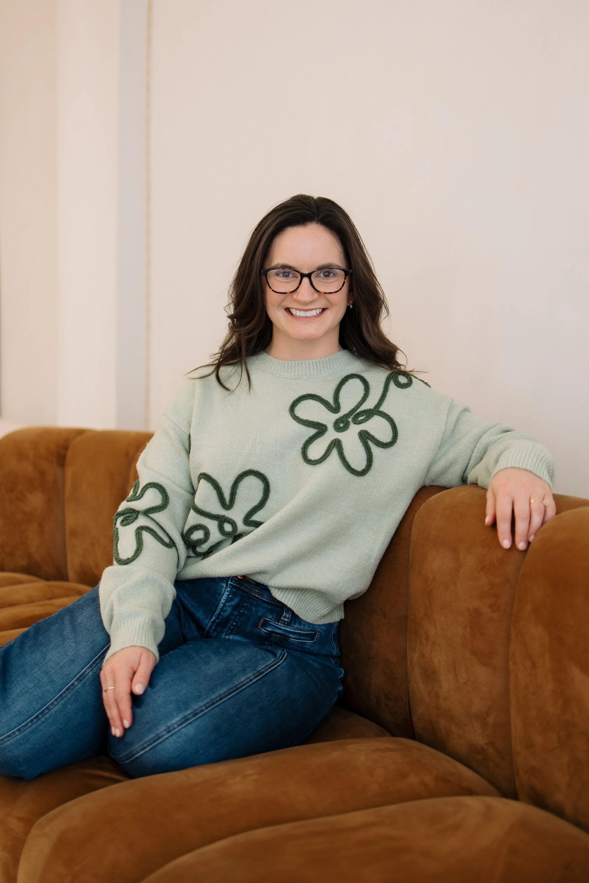 A woman with dark hair, glasses, and a big smile, sitting on a brown couch. She is wearing a light green sweater with dark green floral patterns and blue jeans.