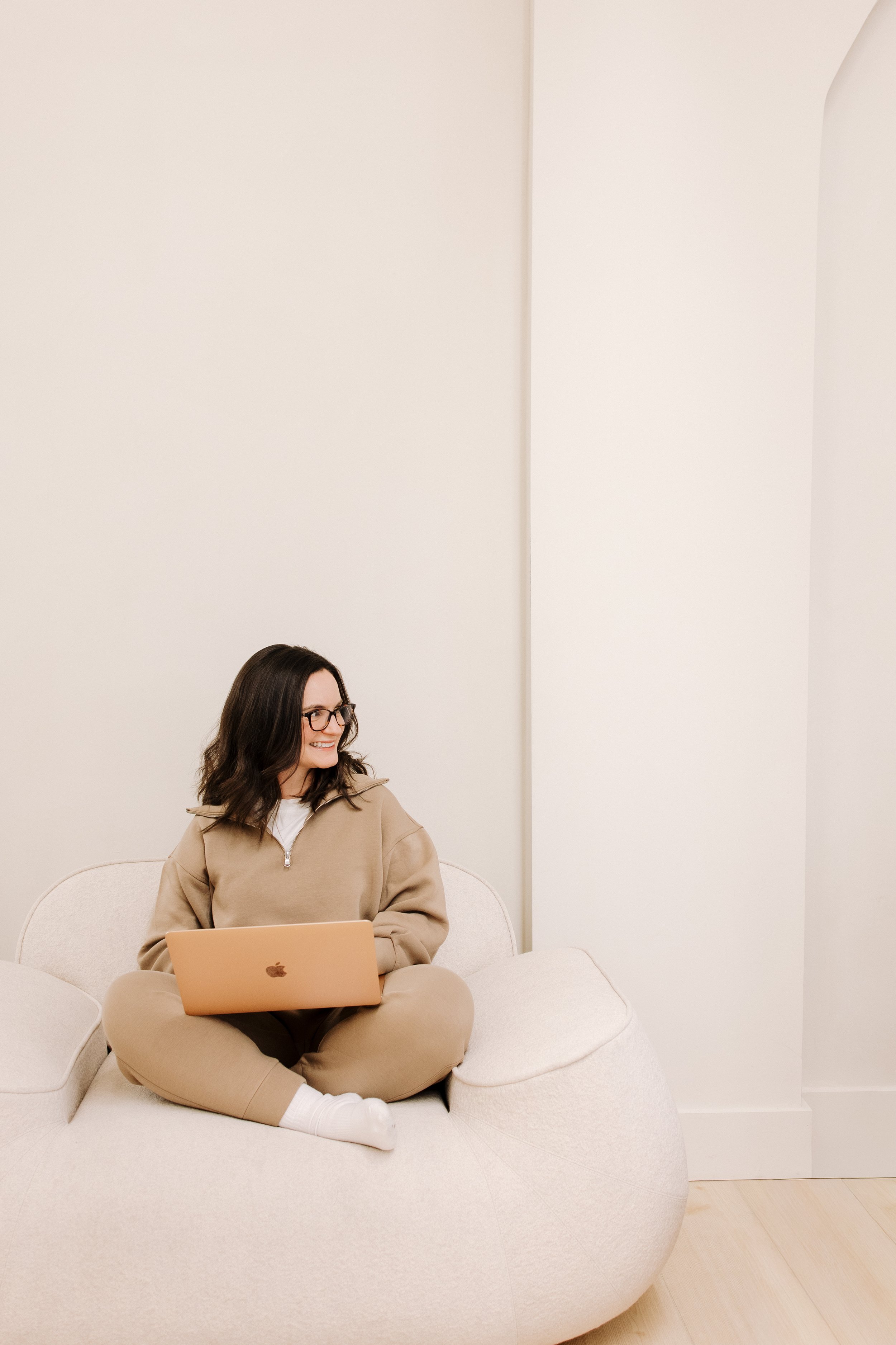 Woman sitting cross-legged on a beige sofa with a laptop, wearing glasses and a beige sweatshirt, smiling and looking to the right in a minimalistic room with white walls.