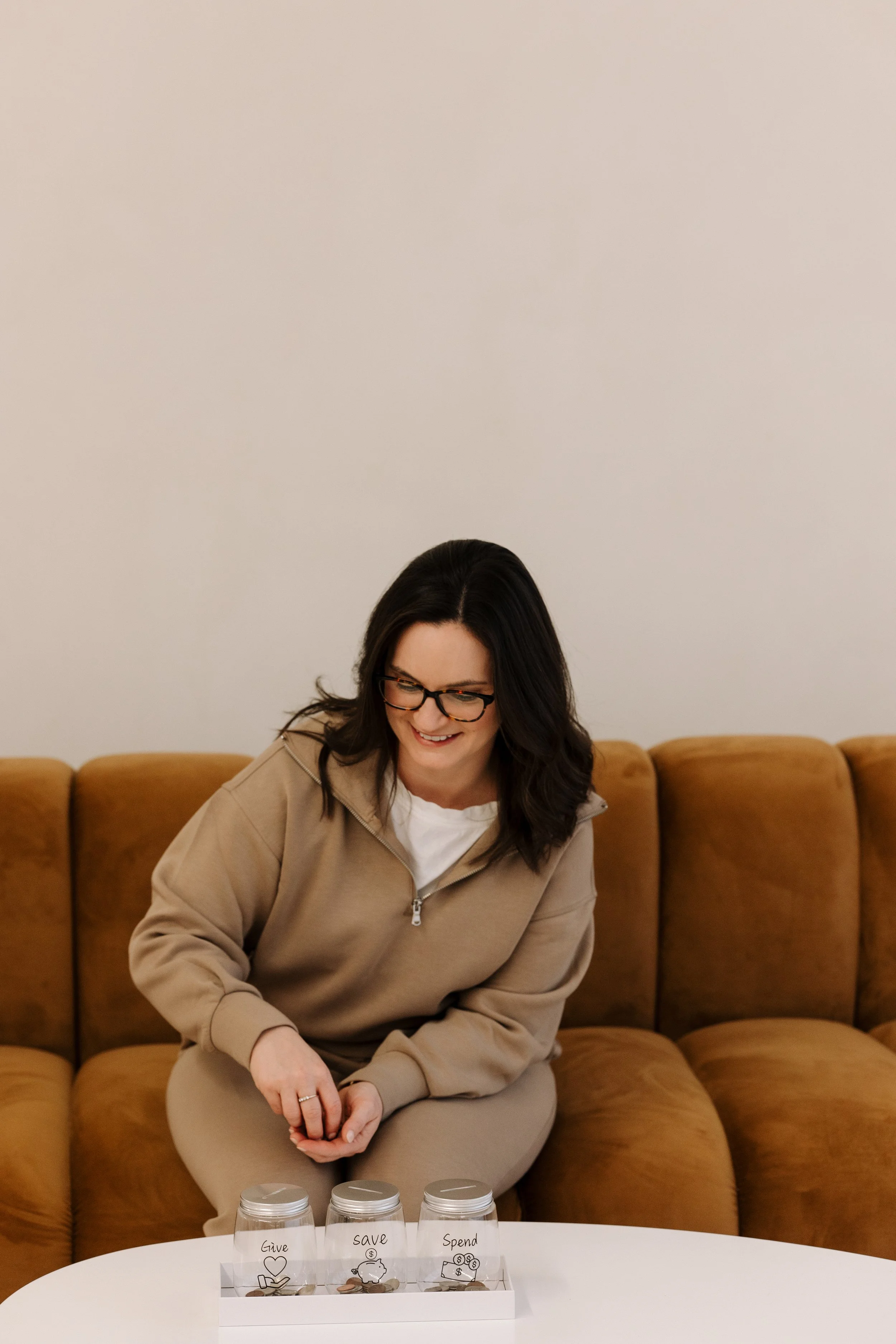 Woman with glasses sitting on a brown sofa, smiling, with a white table in front of her holding three glass jars labeled 'Give', 'Save', and 'Spend' with coins inside.