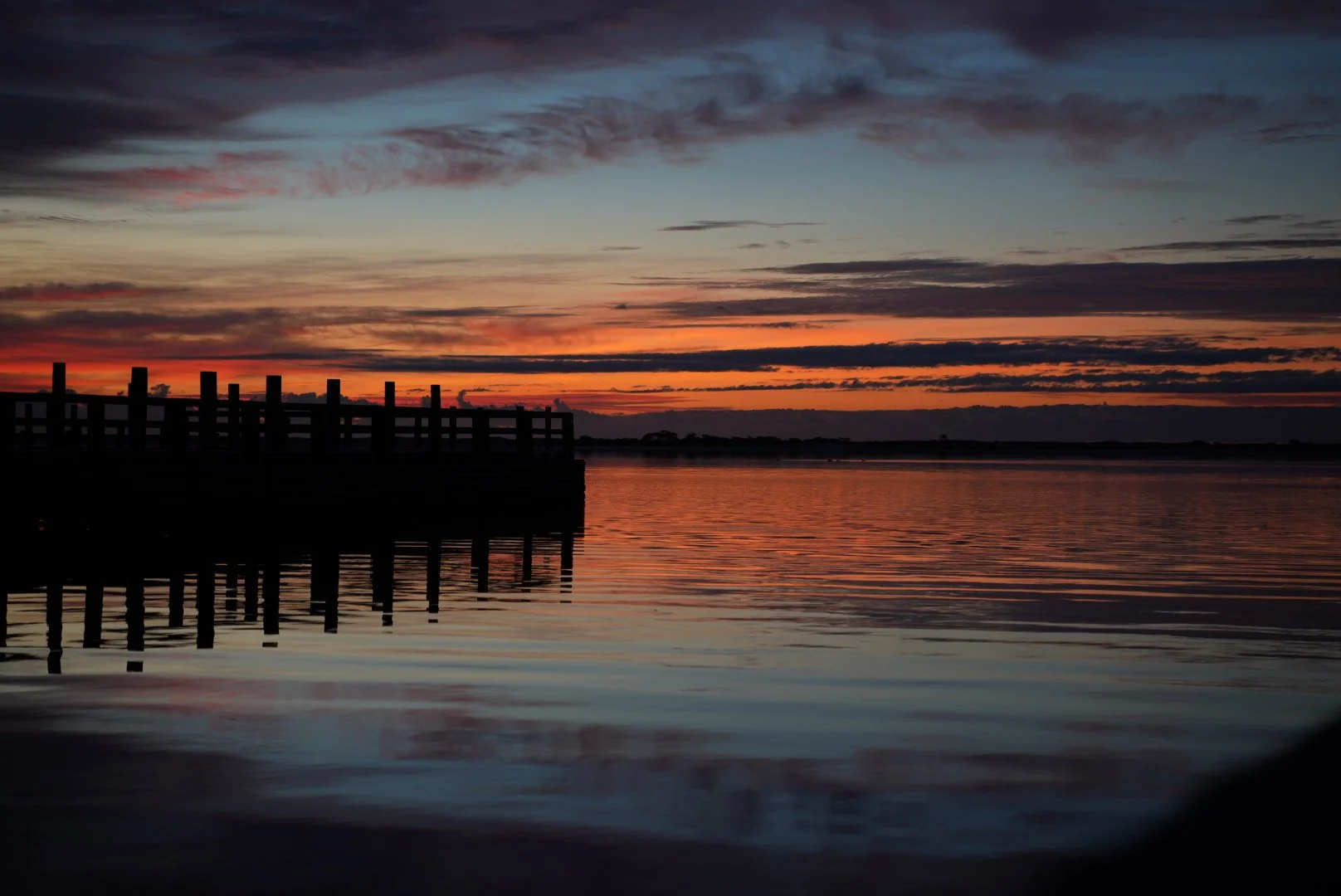 Sunset over a calm body of water with a silhouetted pier on the left side, colorful clouds in the sky.