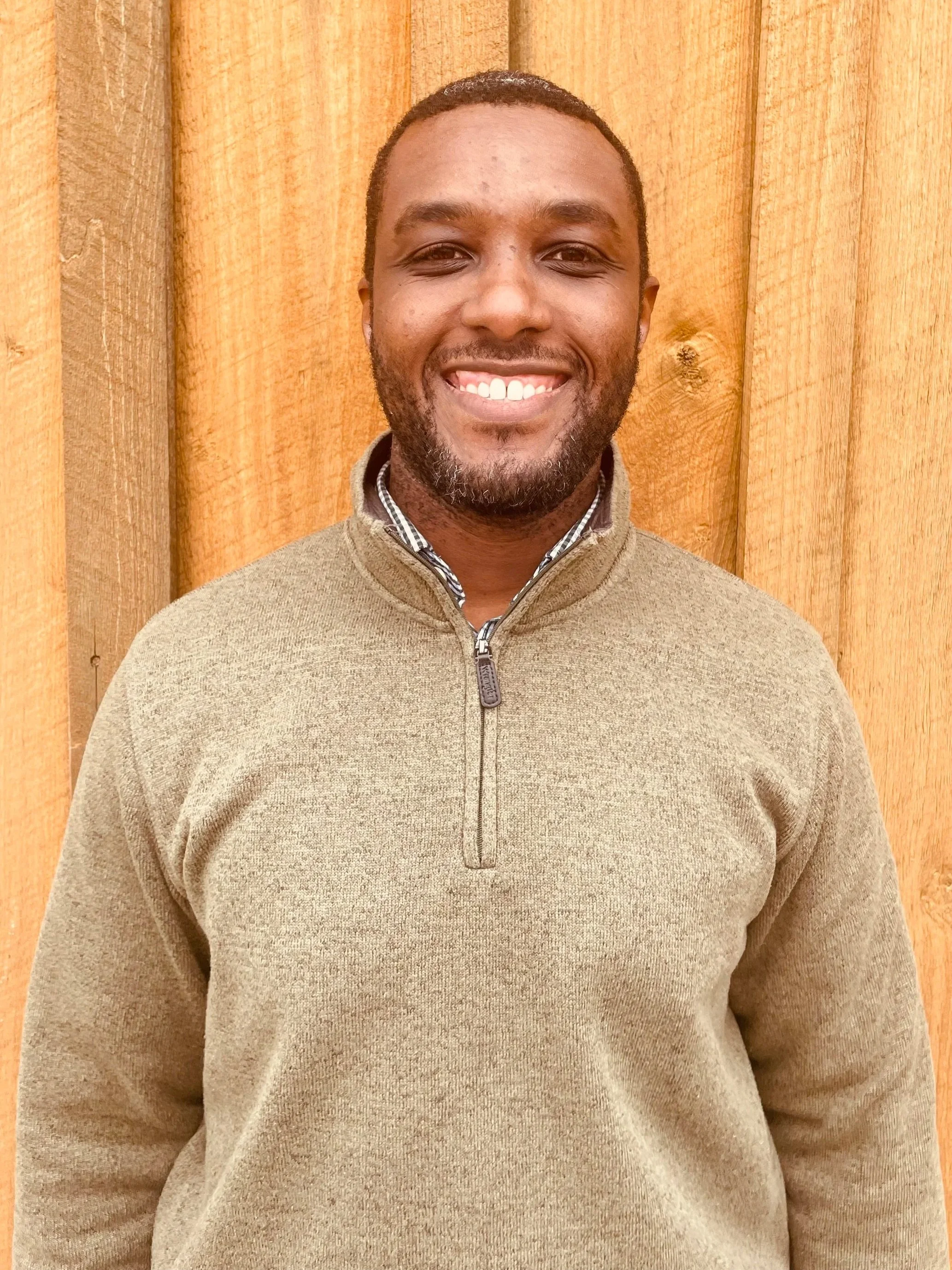 A smiling man standing in front of a wooden background, wearing a beige zip-up sweater.