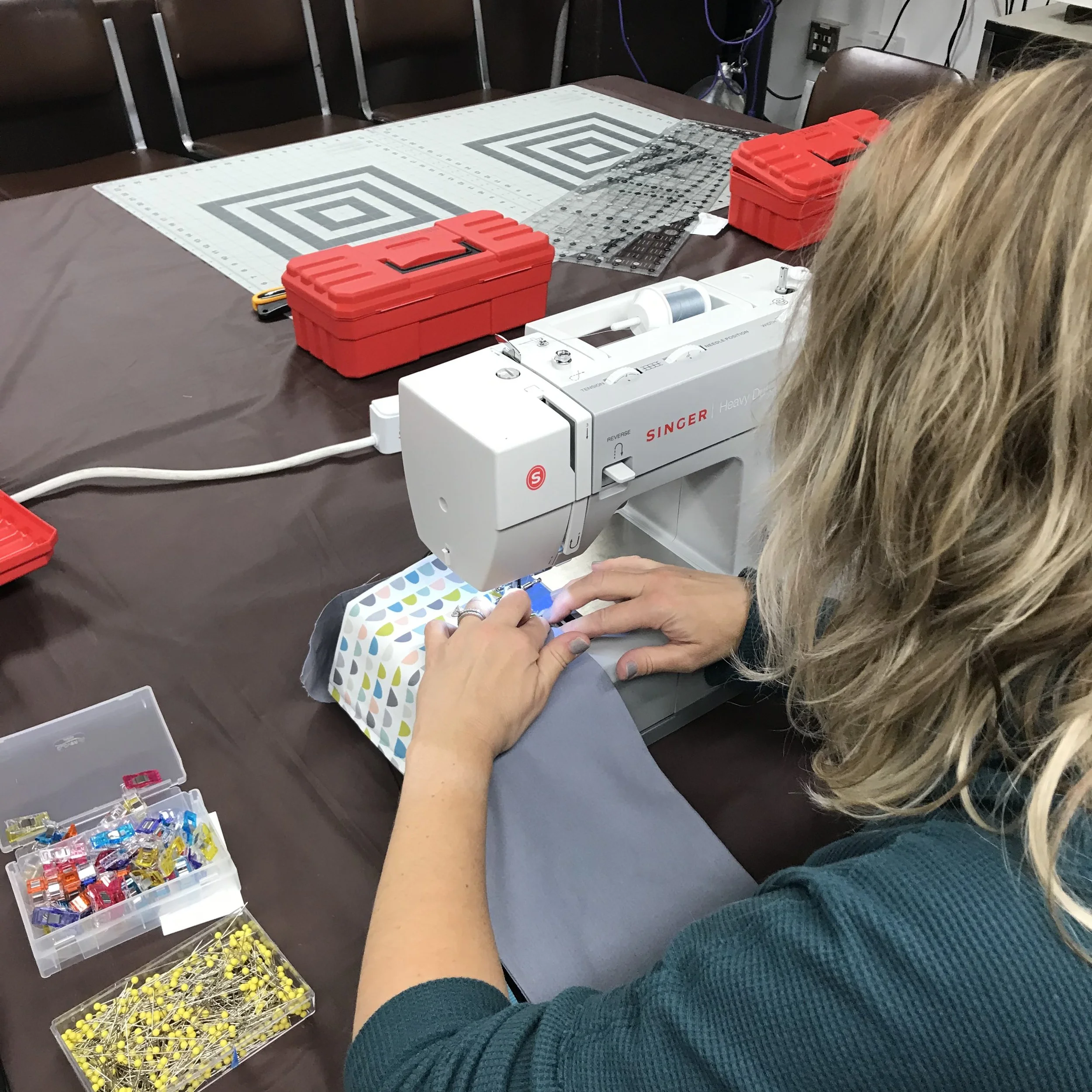 A woman sewing fabric on a sewing machine in a workshop. The table has sewing supplies including boxes of pins and clips.