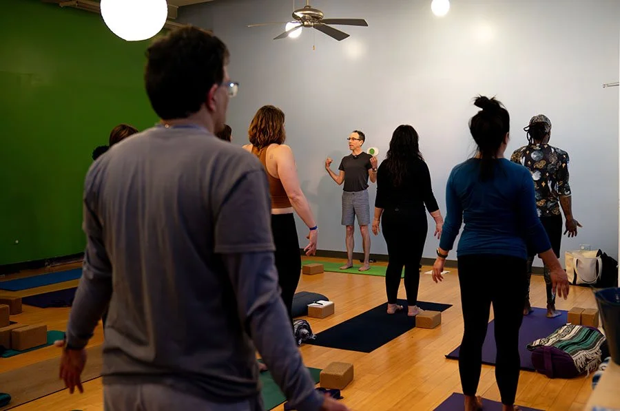 People participating in a yoga class in a studio, some standing on yoga mats, with the instructor, Adam Lazar, at the front, green wall on the left, white wall on the right, ceiling fan, and soft lighting.