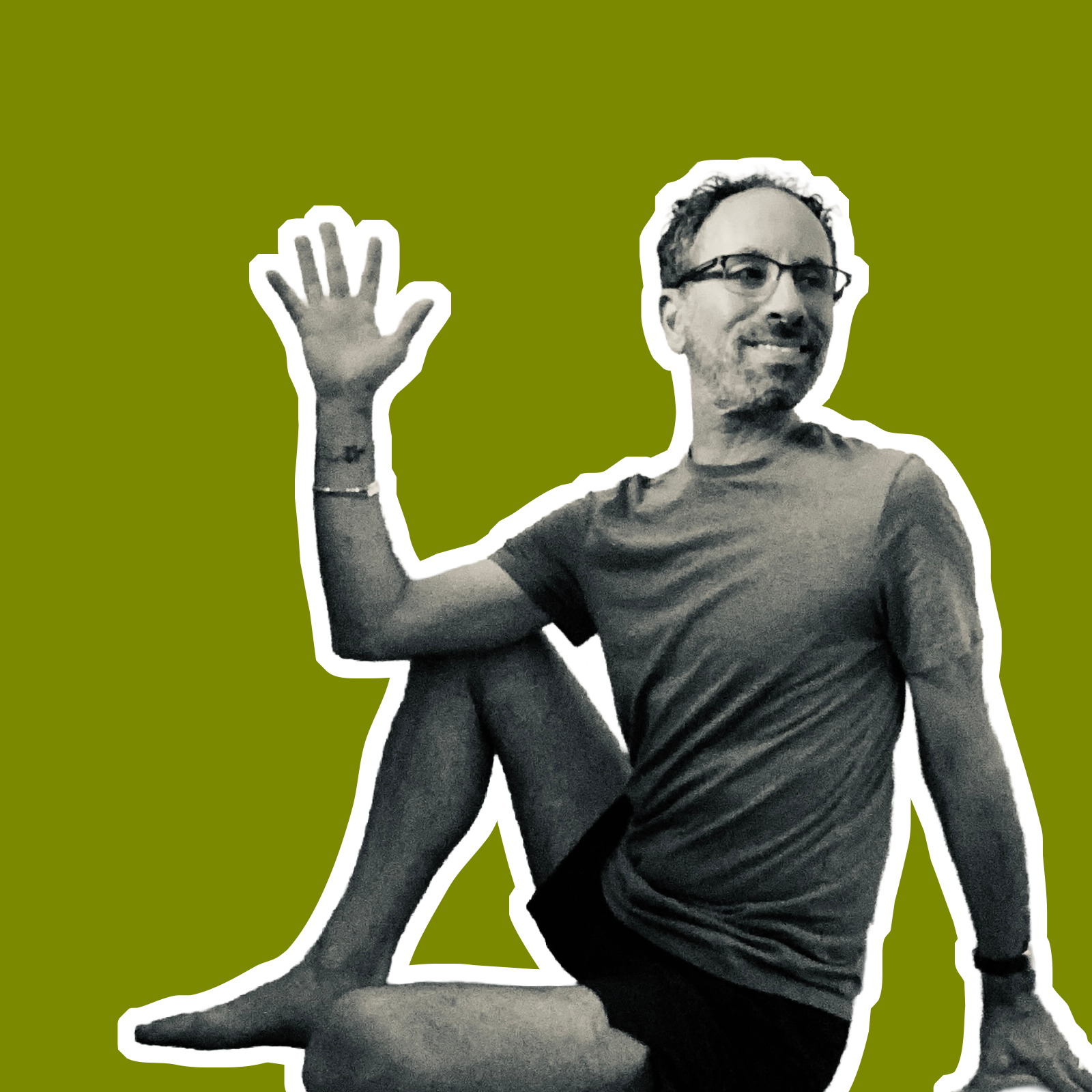 A man sitting cross-legged, smiling, wearing glasses and a t-shirt, waving with his right hand, with tattoos on his left arm, against a solid green background.