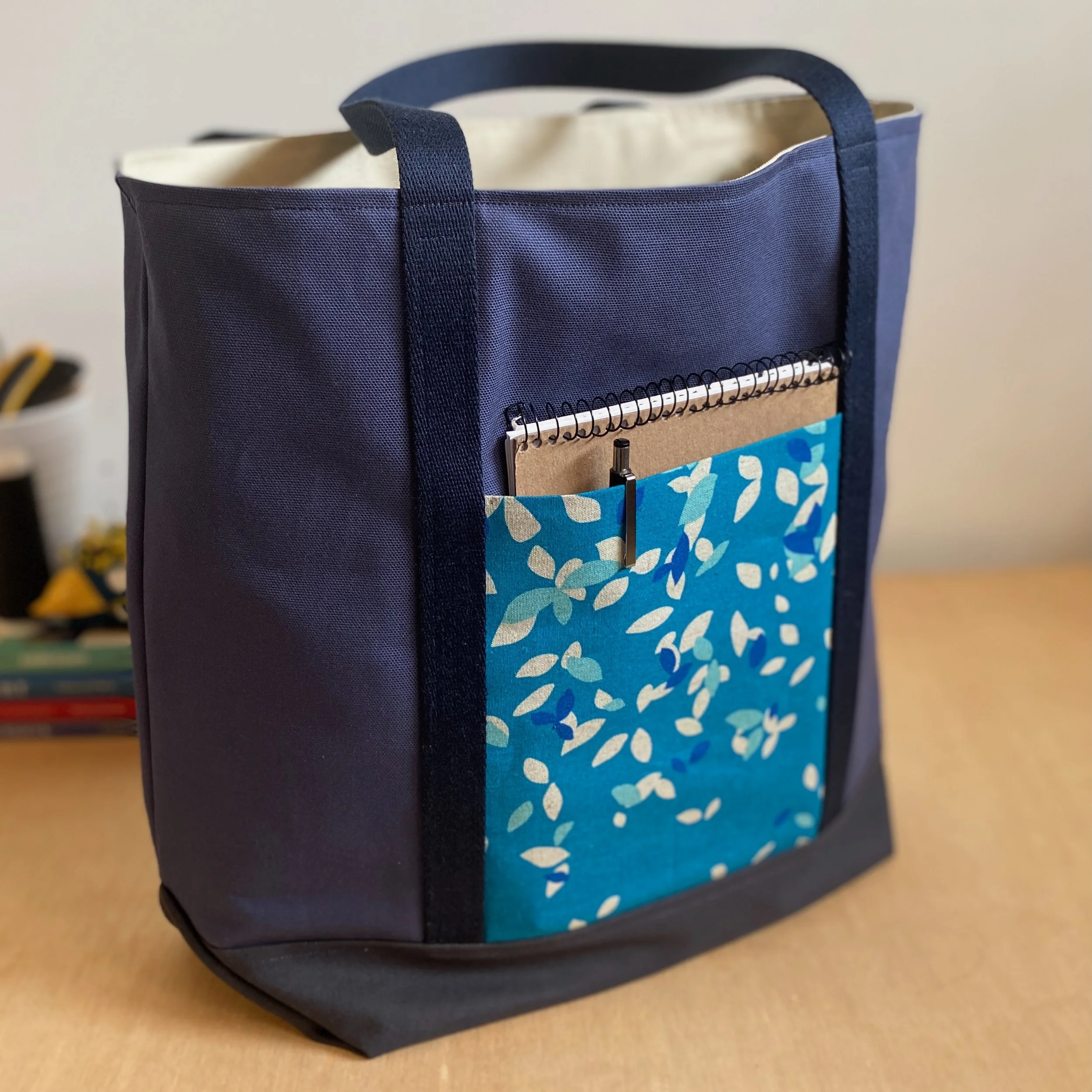 A navy blue tote bag with black straps, containing a notebook, a pen, and a blue patterned pouch, placed on a wooden surface.
