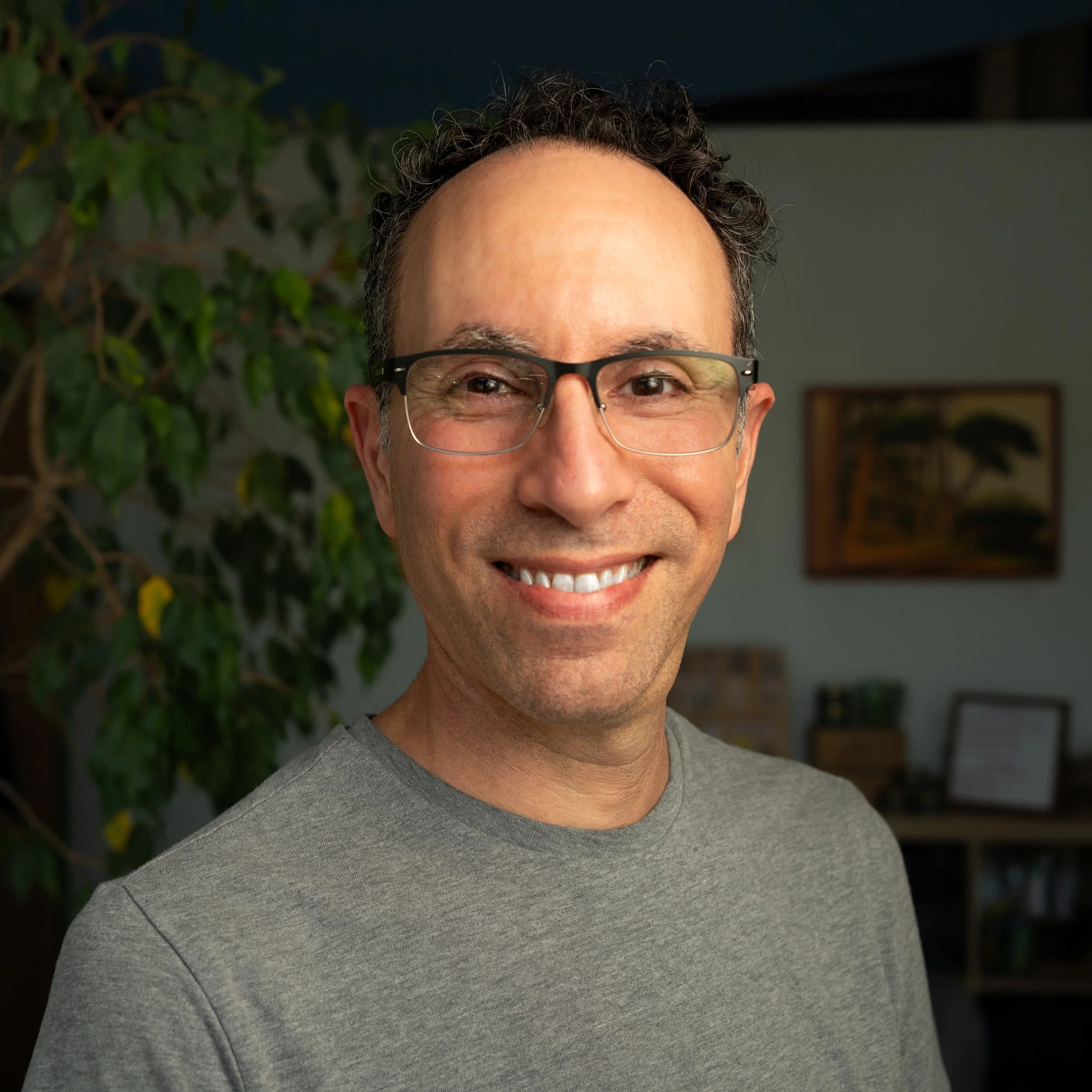 A man, Adam Lazar, with glasses smiling in a room with a large plant and framed pictures on the wall.