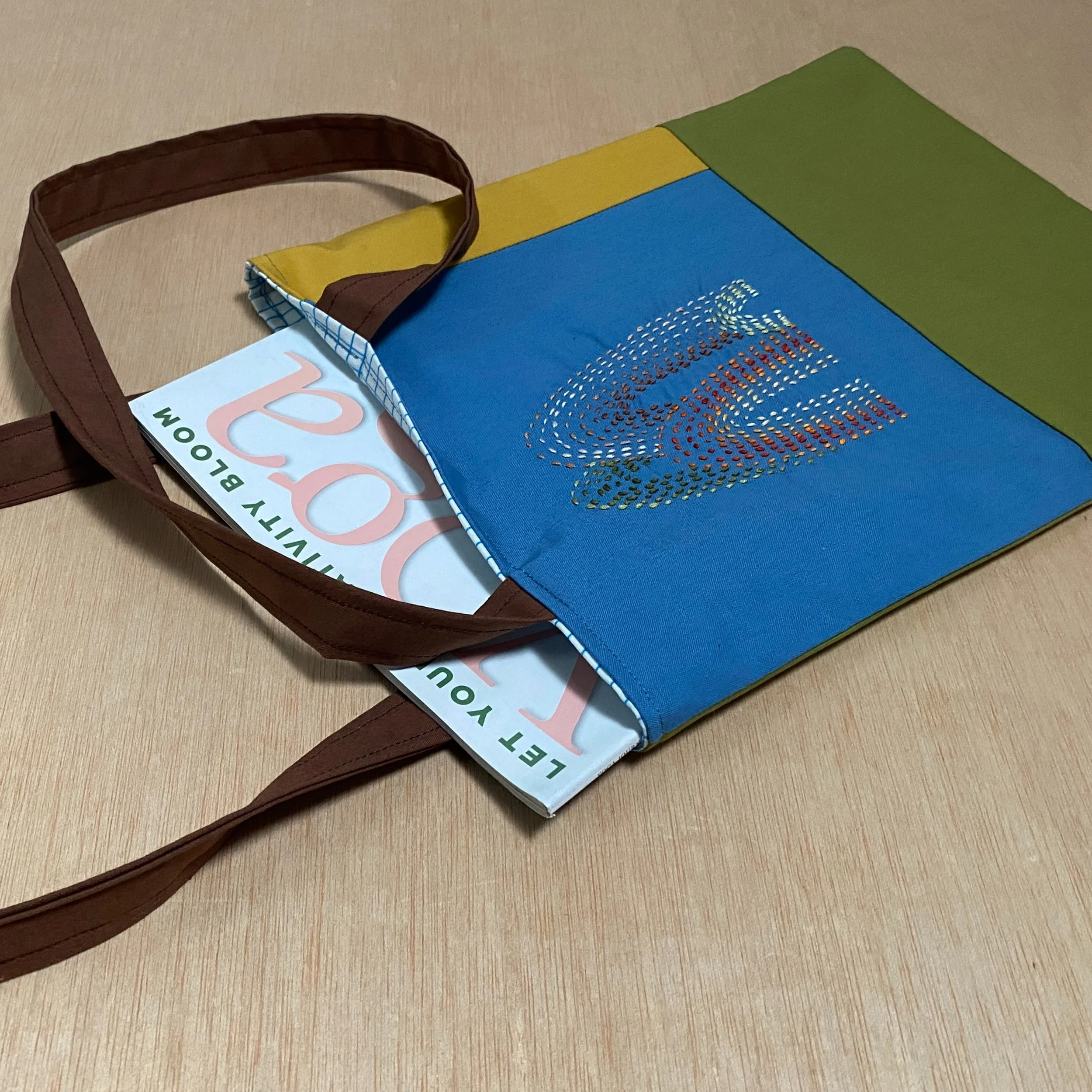 A colorful tote bag with brown handles partially open on a wooden surface, revealing a newsletter or magazine inside.