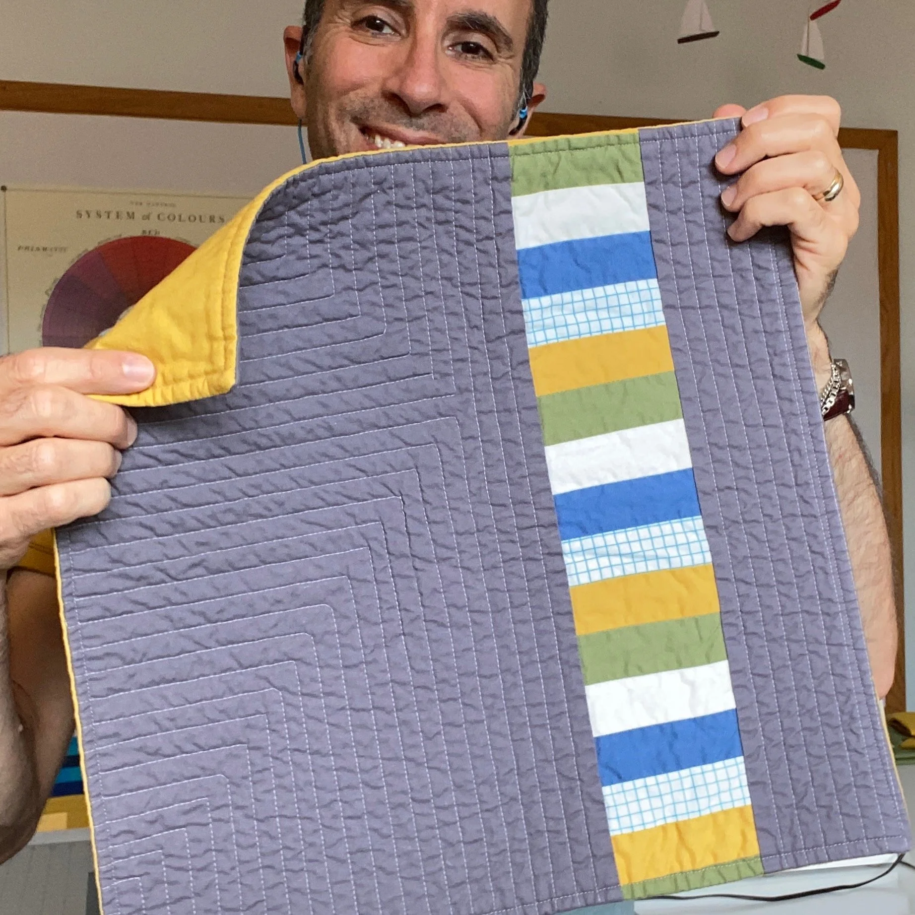 A man smiling and holding up a quilt with a colorful vertical stripe pattern, revealing the quilt's backing.