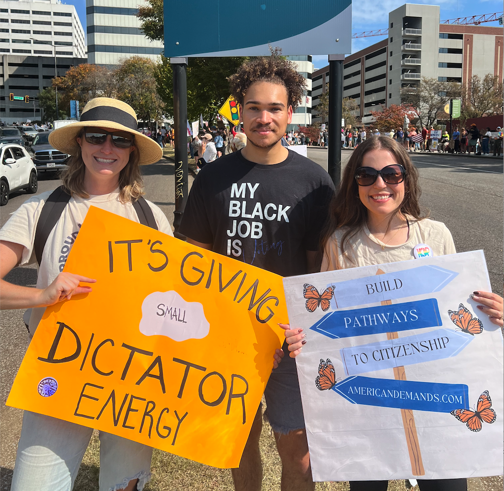 Three people standing outdoors during a protest or march, holding signs. The woman on the left is wearing a straw hat and sunglasses, holding an orange sign that reads 'IT'S GIVING SMALL DICTATOR ENERGY.' The man in the middle is wearing a T-shirt that says 'MY BLACK JOB IS,' with the rest of the text not visible. The woman on the right is wearing sunglasses and holding a sign with colorful arrows and butterflies that says 'BUILD PATHWAYS TO CITIZENSHIP' and includes the website 'AMERICANDAMONDS.COM.' In the background, there are buildings, trees, and other protesters.