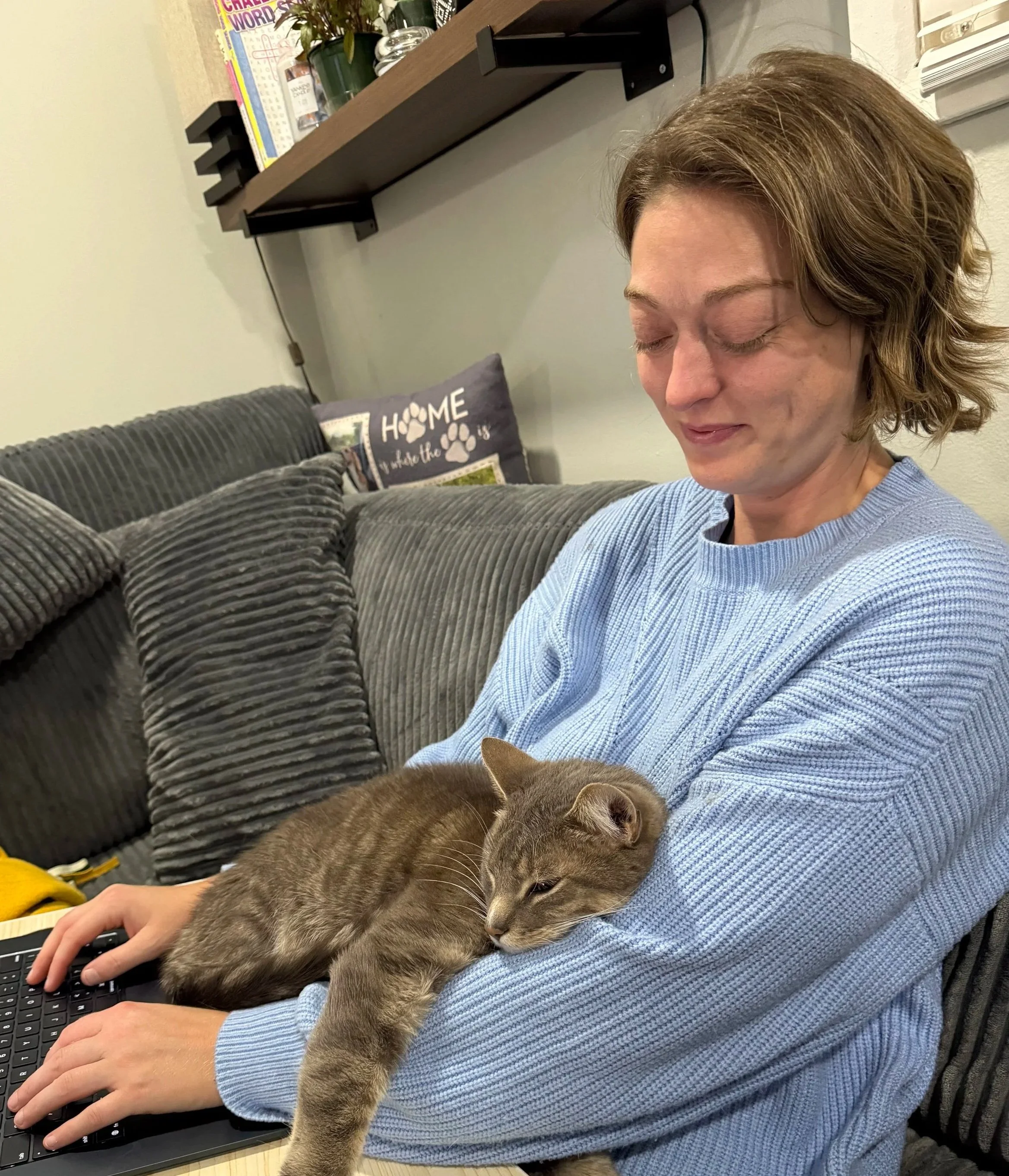 A woman in a blue sweater sitting on a gray couch with a gray tabby cat resting on her lap and leaning on her arm. The woman is smiling with her eyes closed, and the background includes shelves with books and a decorative pillow with the words 'Home is where the paw is' and paw print designs.