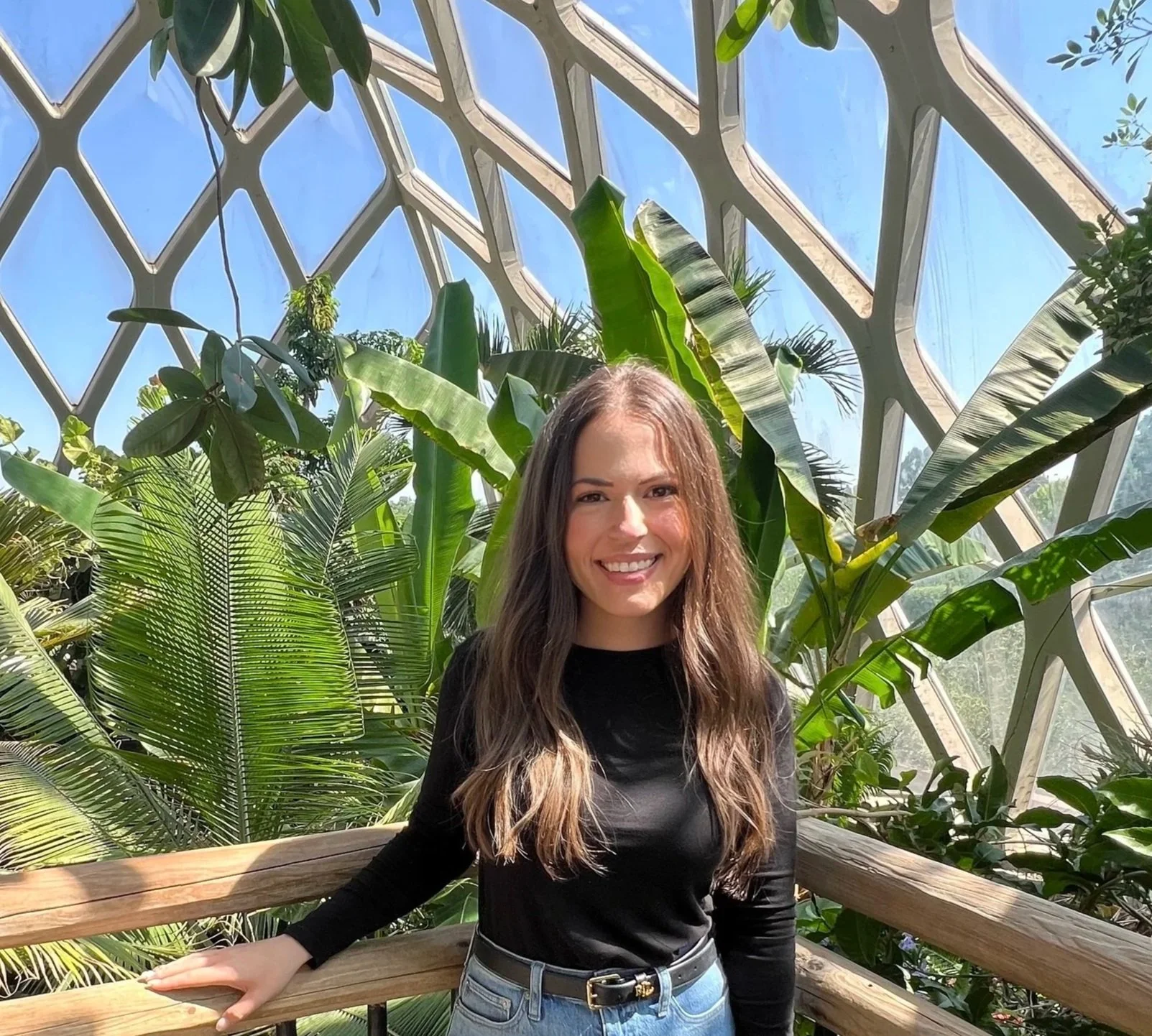 A smiling young woman in black top and jeans standing on a wooden railing inside a glass conservatory with lush green tropical plants and a bright blue sky visible through the glass ceiling.