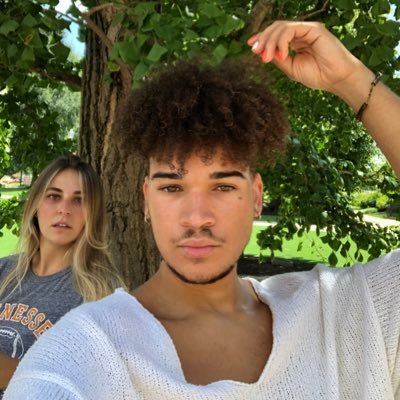 A young man with curly hair taking a selfie outdoors under a tree, with a young woman in the background.
