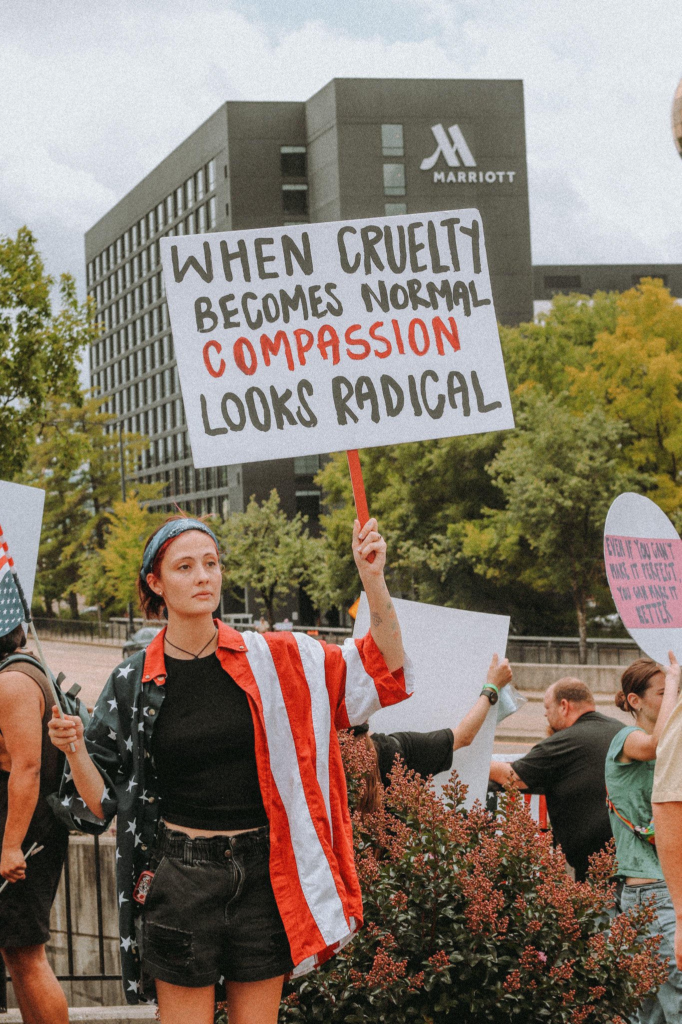 A young woman holding a protest sign that reads 'When cruelty becomes normal, compassion looks radical.' She is wearing a shirt with an American flag design and a National flag headscarf, standing at a rally or protest with other people, with trees and a Marriott hotel building in the background.