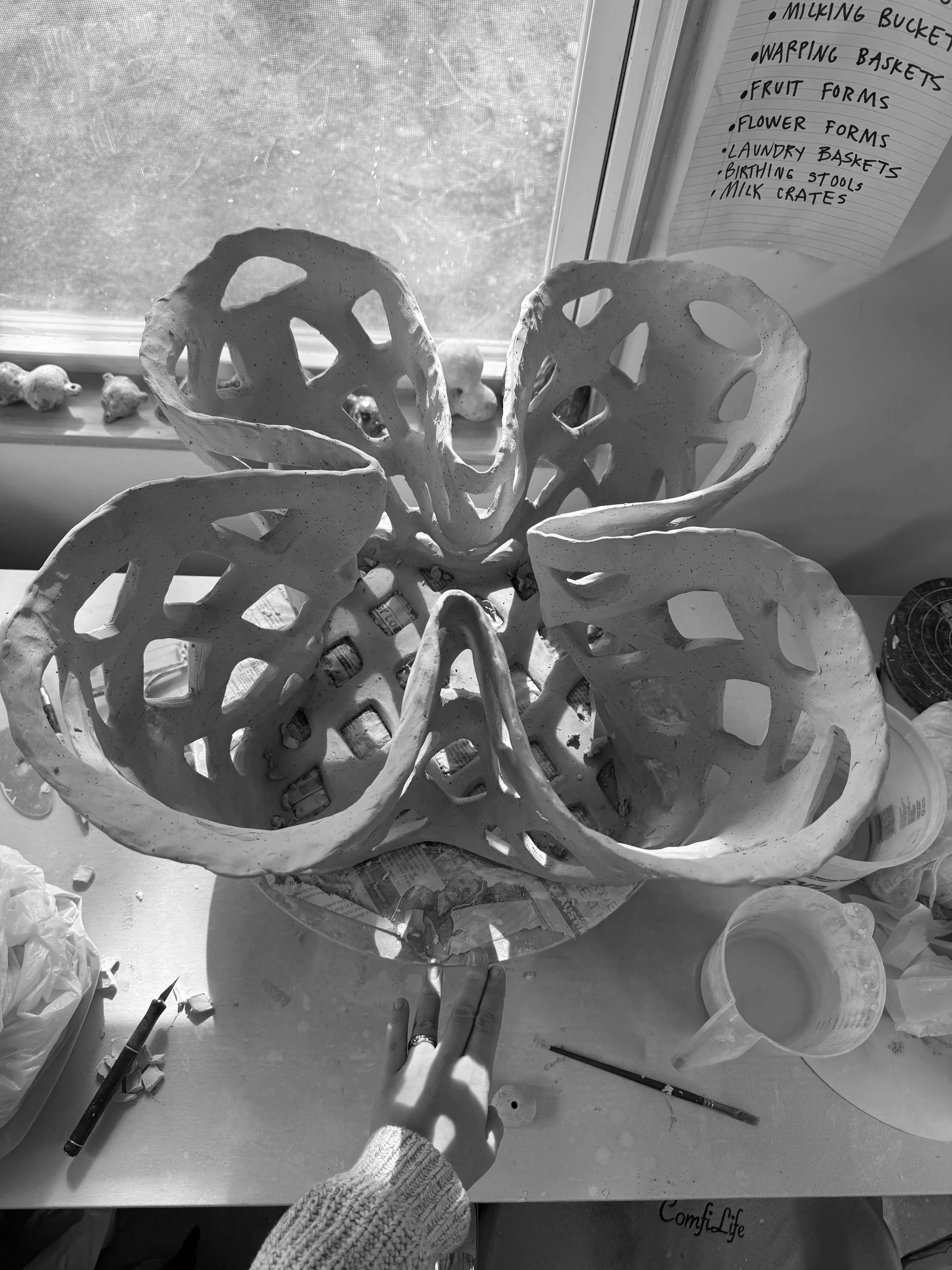 A ceramic butterfly sculpture with openwork details on a table, surrounded by art supplies including paintbrushes, a cup of water, and paper. In the background, a window with a list of tasks or materials written on a notepad is visible.