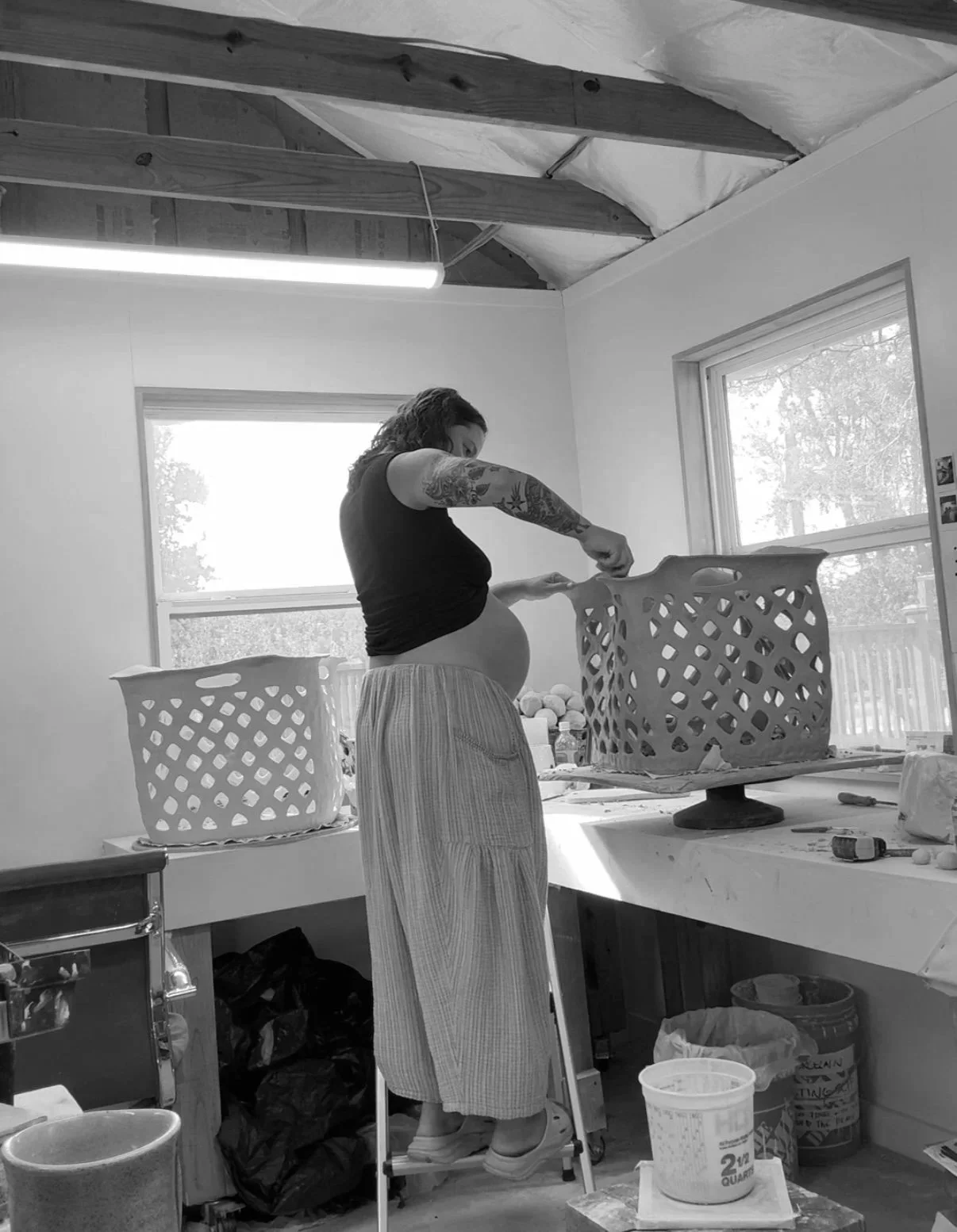 A pregnant woman with tattoos is working on a large basket sculpture in a studio space with unfinished ceiling and windows, standing on a small ladder.