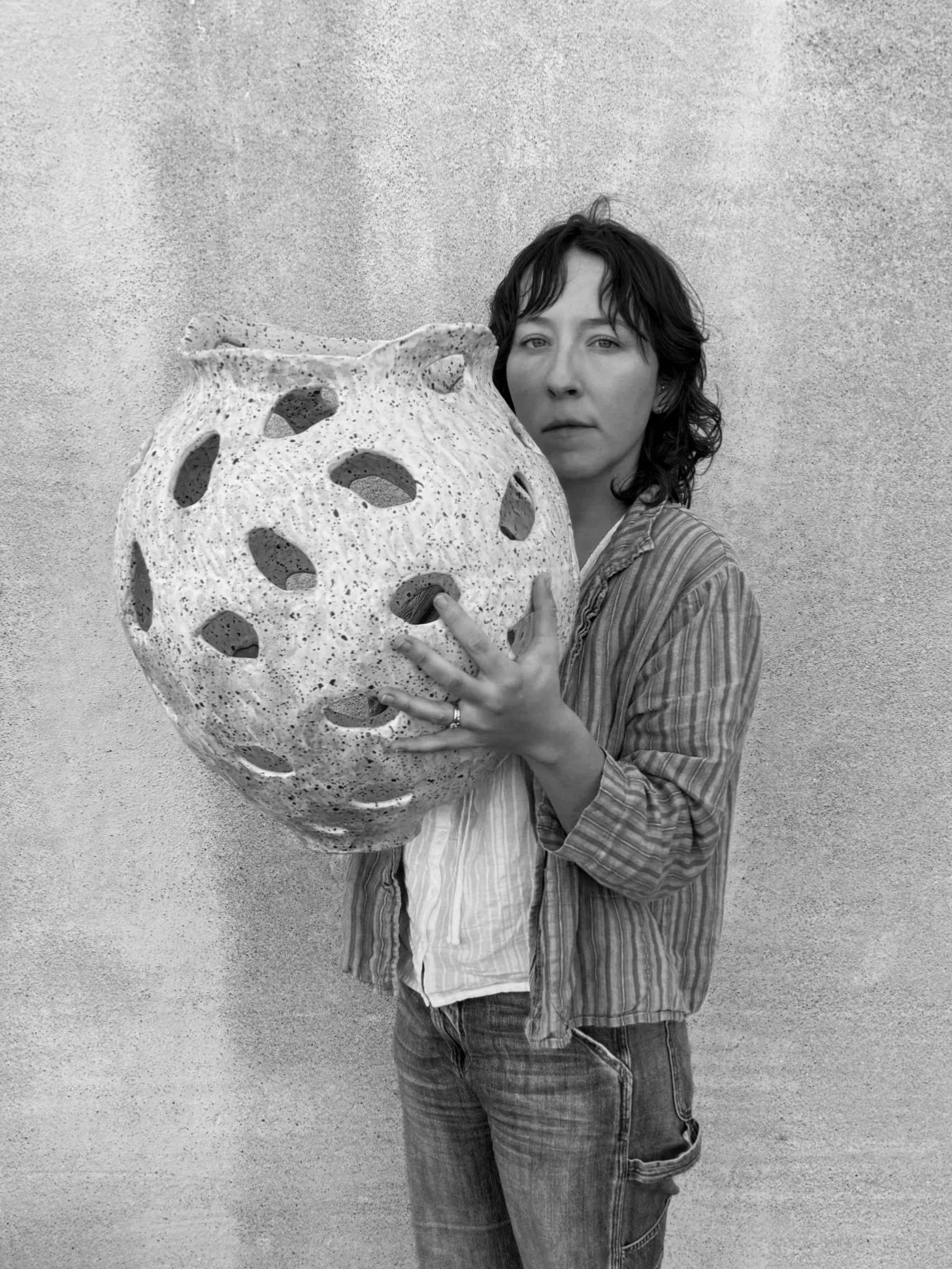 A woman with short, wavy hair standing against a plain wall, holding a large, textured spherical object with multiple holes.
