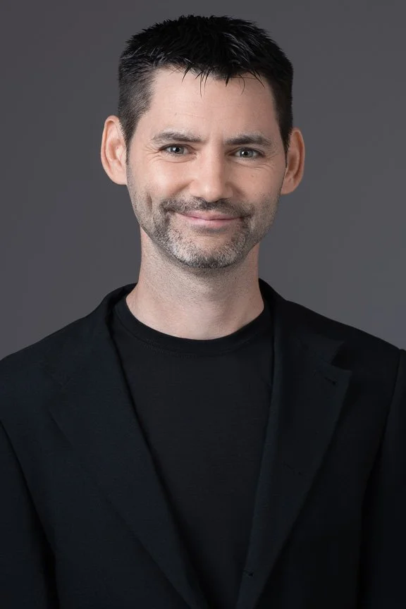 Portrait of a man with short dark hair, slight beard, wearing a black blazer and black shirt, smiling against a gray background.