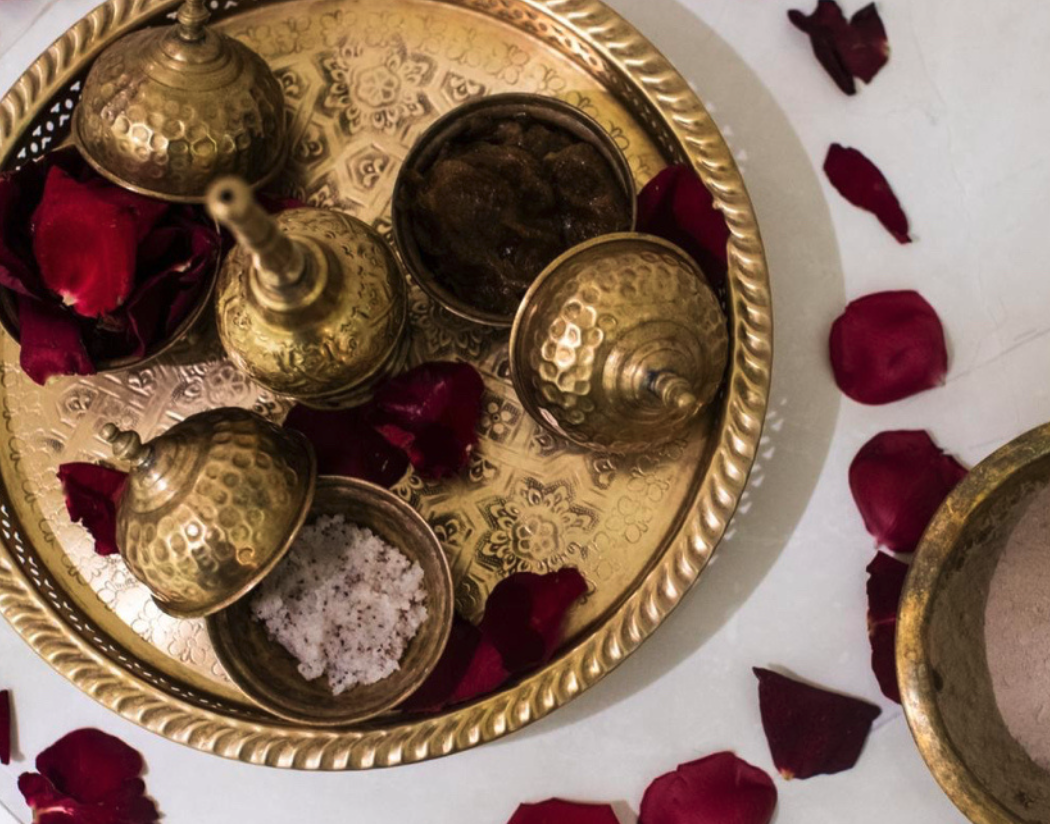 Plate ronde en or avec plusieurs contenants pour épices ou huiles, entouré de pétales de rose rouges, sur un fond blanc.