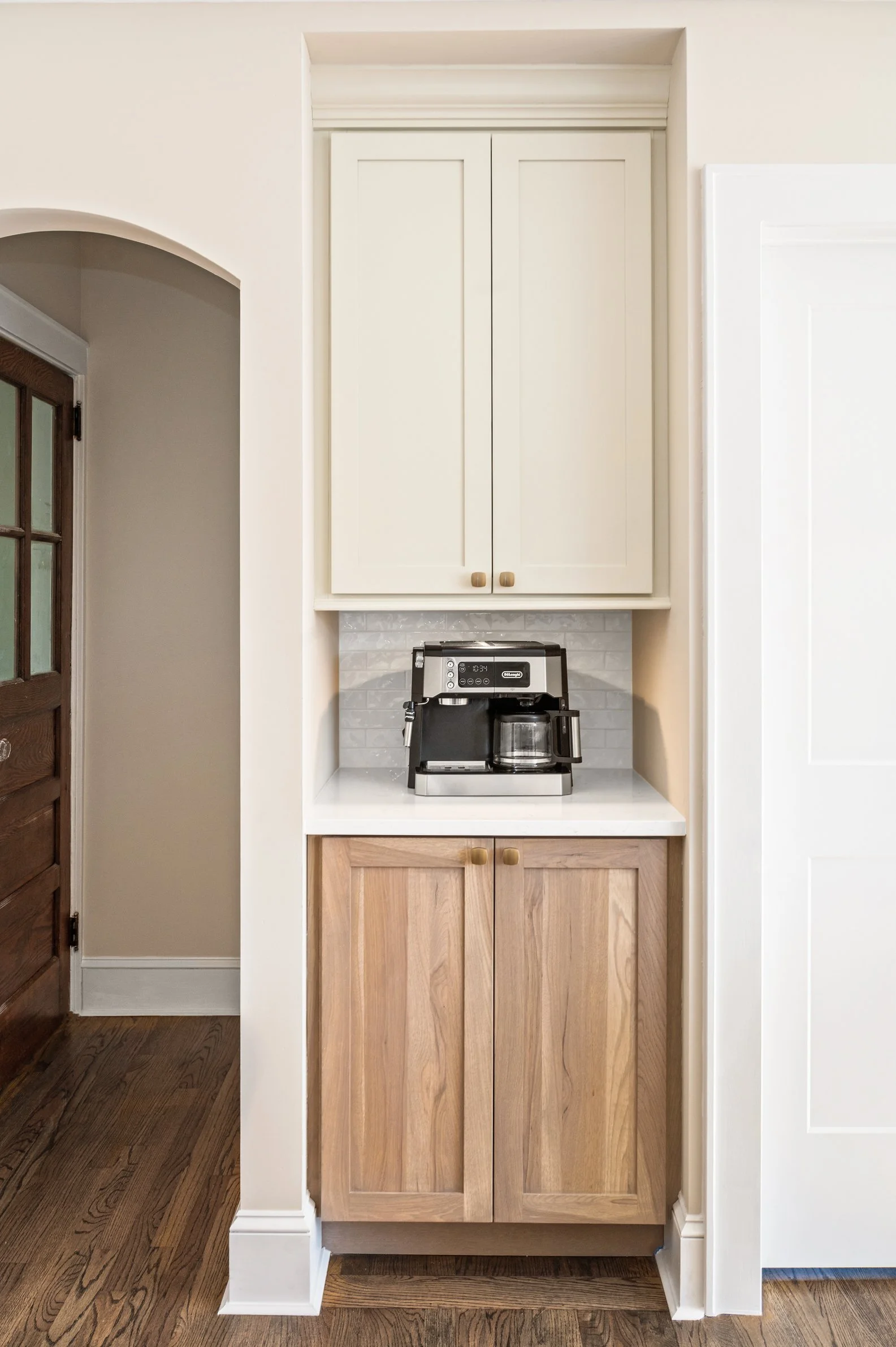 Coffee maker on a kitchen countertop between two cabinets, with a backsplash and hardwood floor.