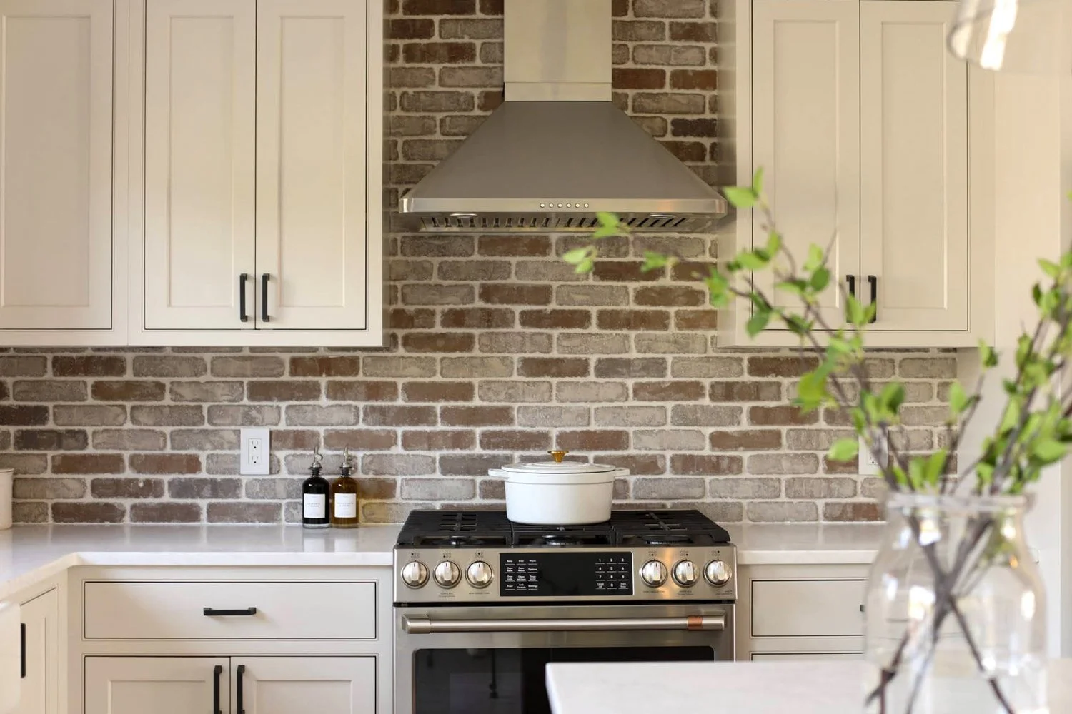 White kitchen with brick wall backsplash, stainless steel stove with white pot on top, white cabinets with black handles, a vase with green plant on counter, and oil bottles nearby.
