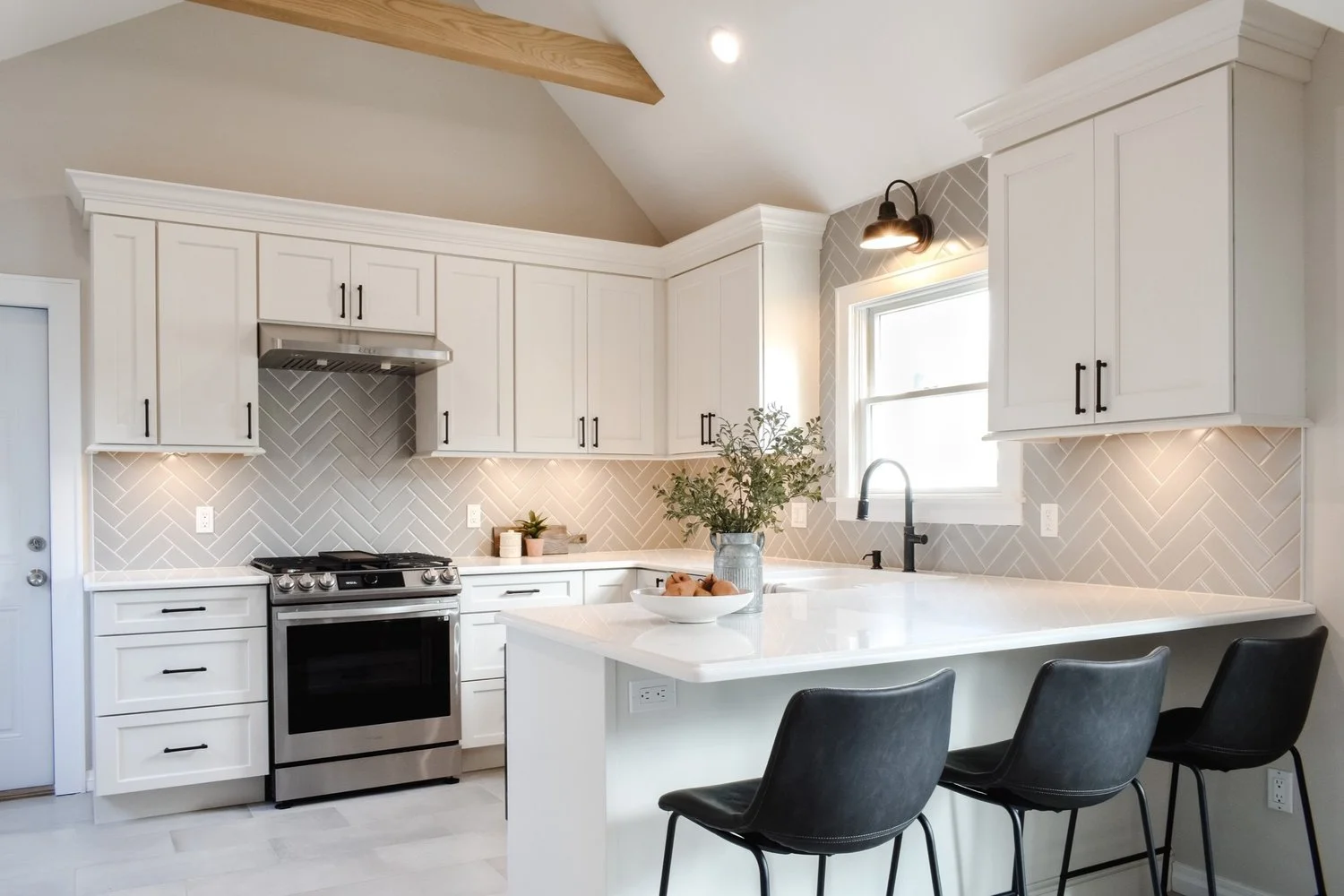 Modern white kitchen with grey herringbone backsplash, black cabinet handles, black chairs at island, stainless steel stove, and a window above the sink.