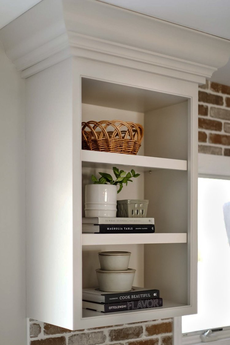 White kitchen cabinet with bowls, books, potted plant, and woven basket inside, next to a brick wall.