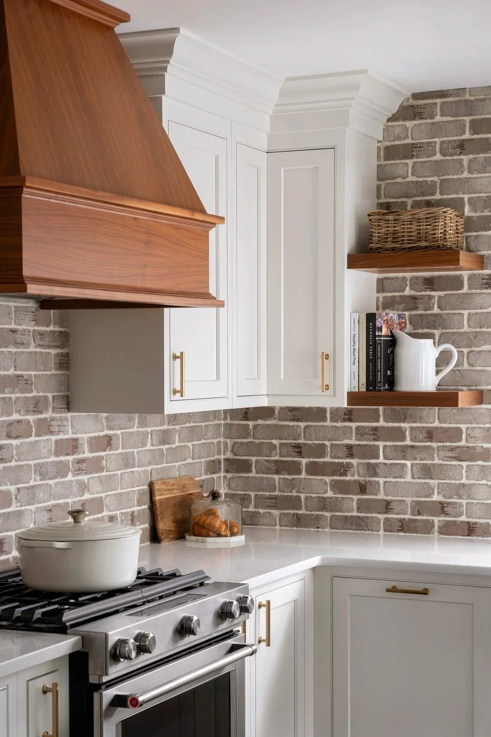 Kitchen corner with white cabinets, brick wall, open shelves with woven basket, books, and white pitcher, stainless steel stove with a white pot, and a white countertop with croissants in a glass cloche.