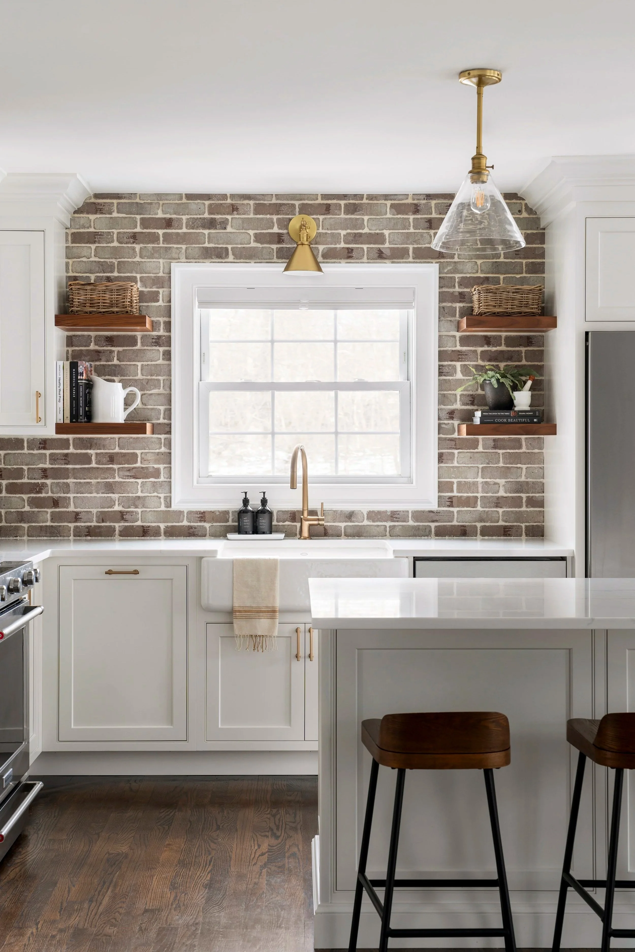 Modern kitchen with white cabinets, brick wall backdrop, window, and wooden shelves with decor and cookbooks.