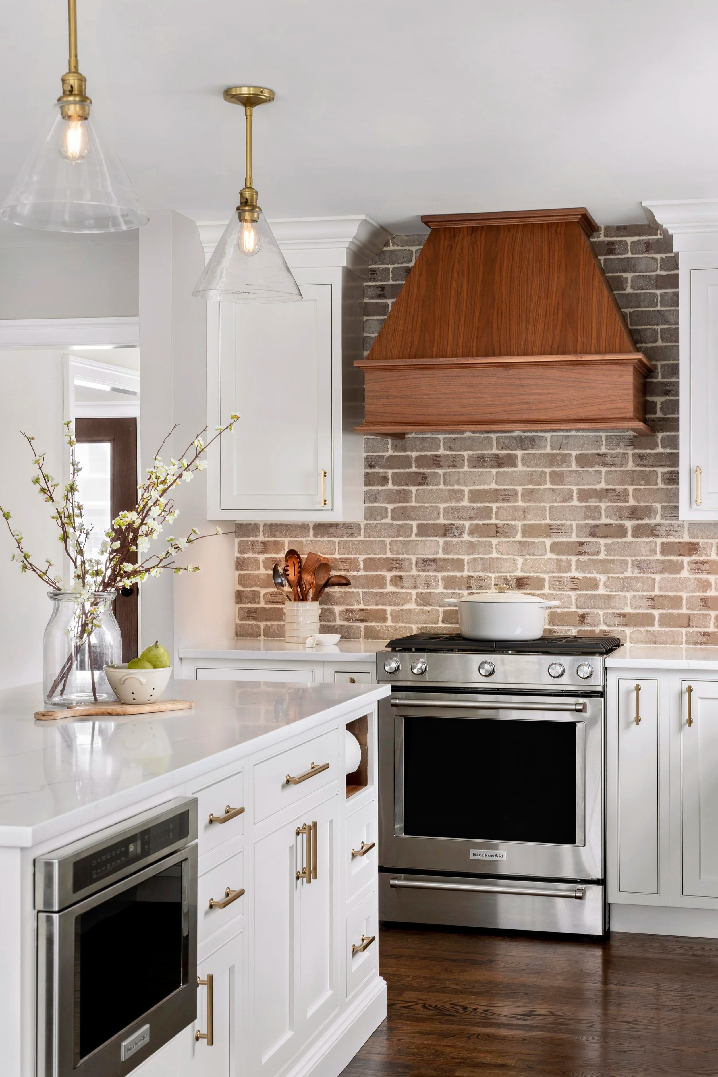 Modern kitchen with white cabinets, a stainless steel oven, a brick wall backsplash, and a wooden range hood. Decor includes a large glass vase with branches, a small bowl of pears, and a utensil holder.