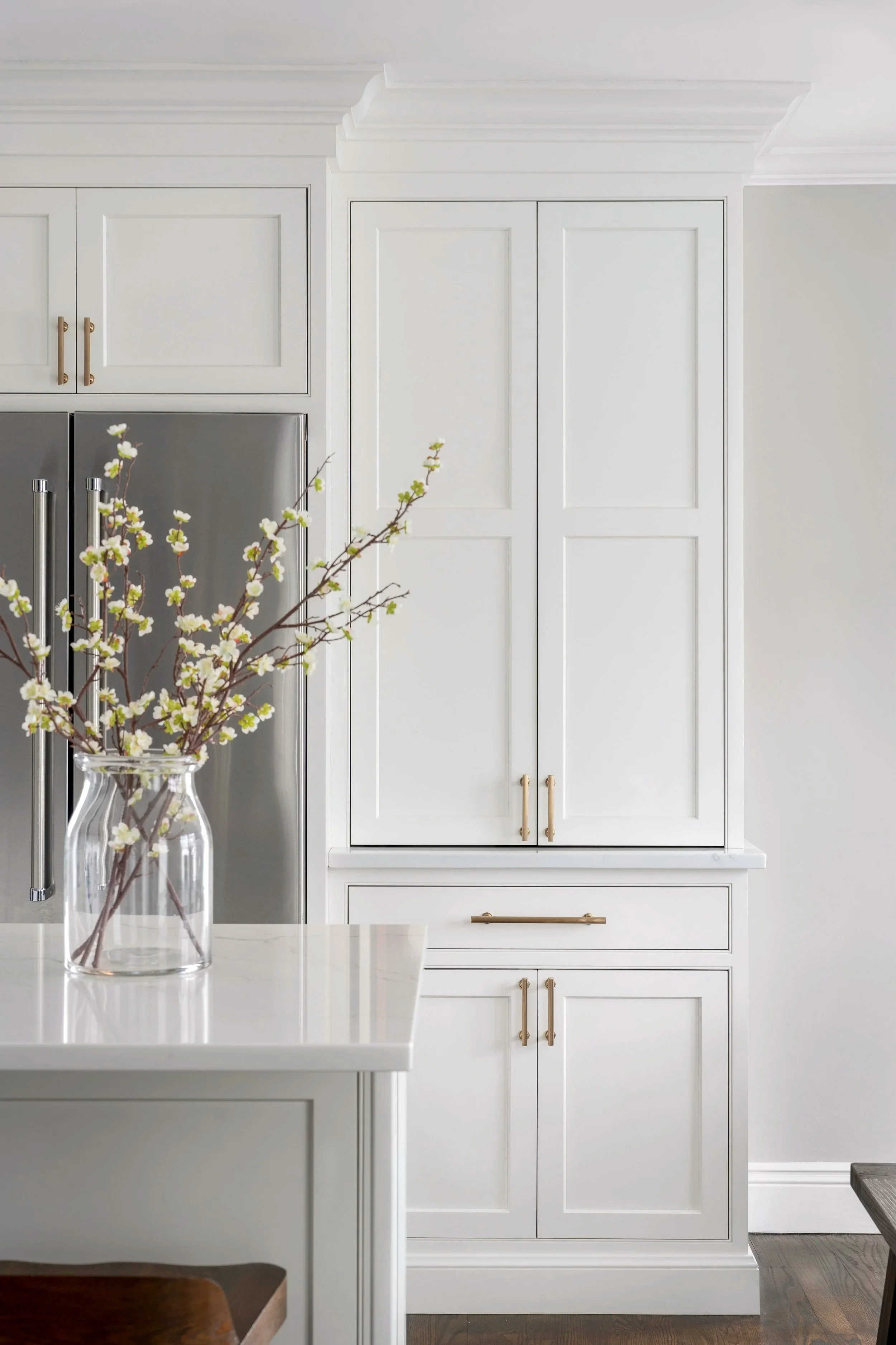 White kitchen cabinet with gold handles, stainless steel refrigerator, and a glass jar with flowering branches on a white countertop.