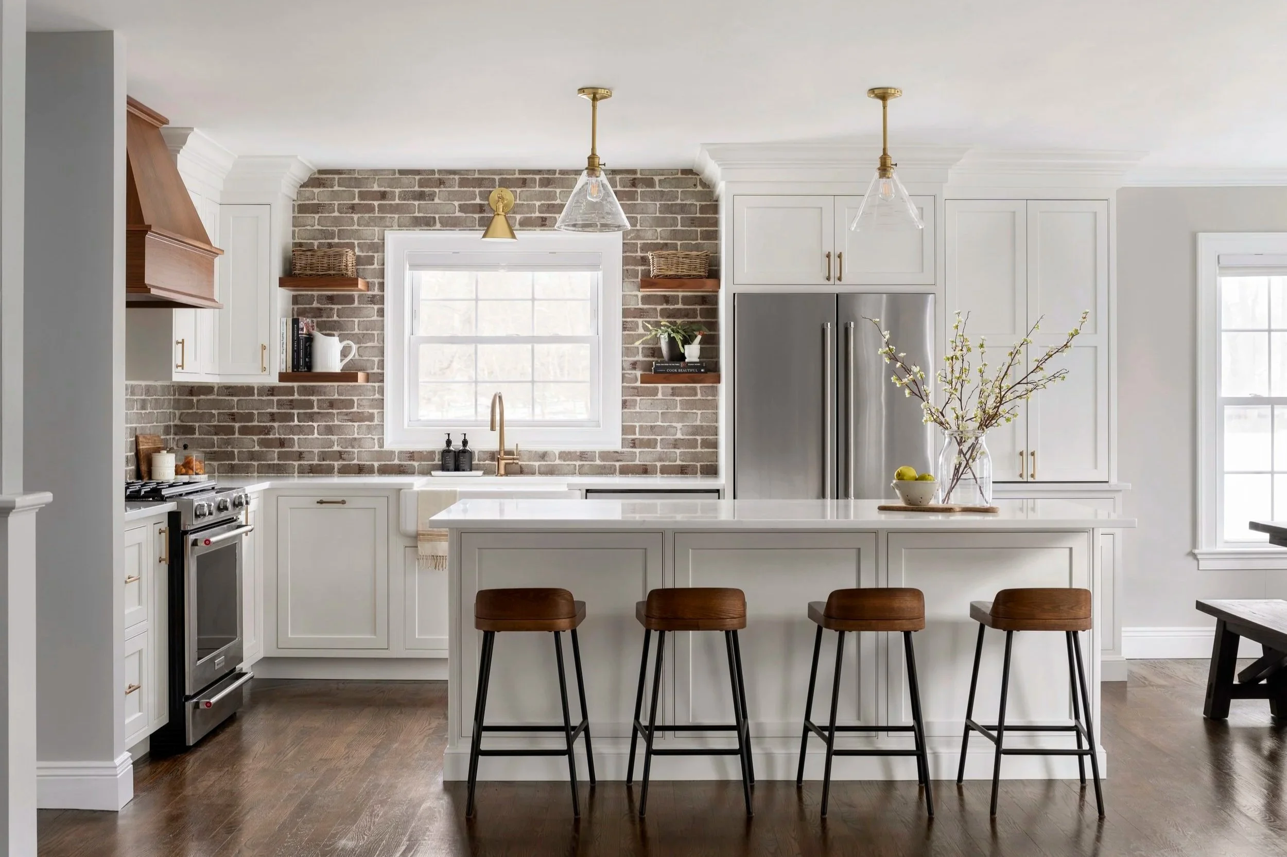 Modern kitchen with white cabinets, a brick backsplash, wooden shelves, stainless steel appliances, a white island with four wooden stools, and large windows letting in natural light.