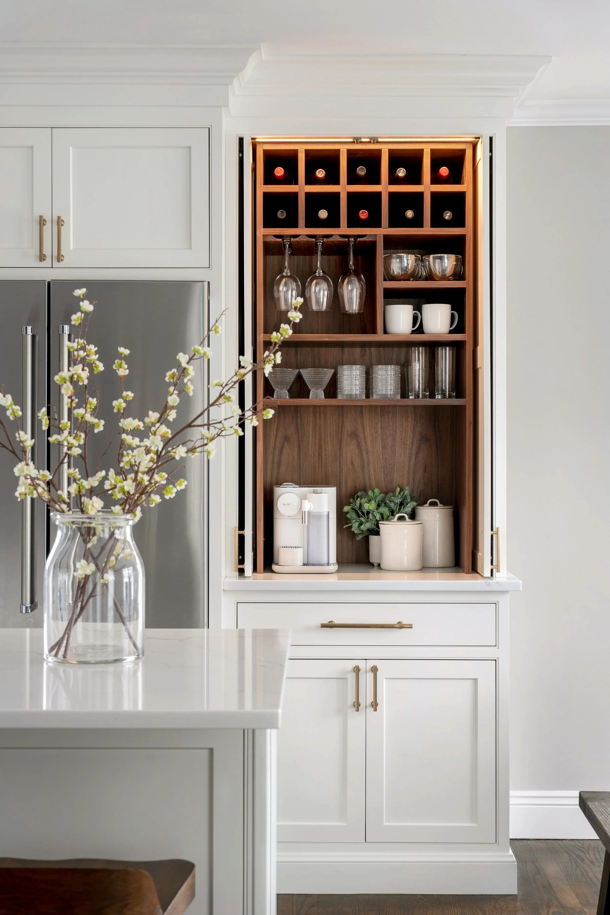 Kitchen cabinet with organized wine bottles, glasses, bowls, mugs, cups, and a coffee maker, with a vase of flowers on the counter.
