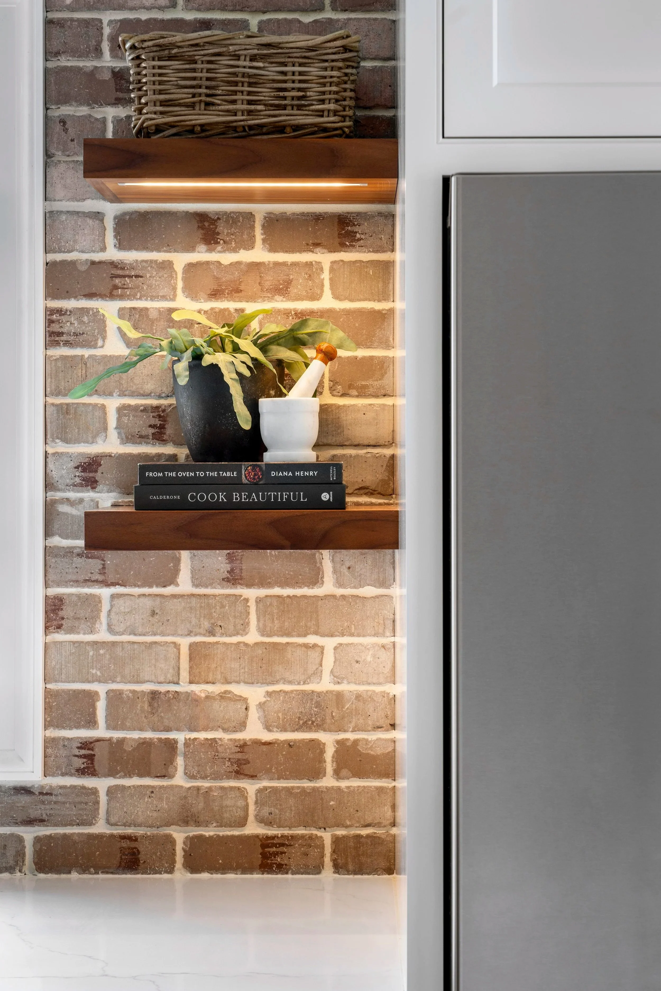 Close-up of a kitchen shelf with potted plant, books, and brick wall background, partially obscured by a white kitchen cabinet.