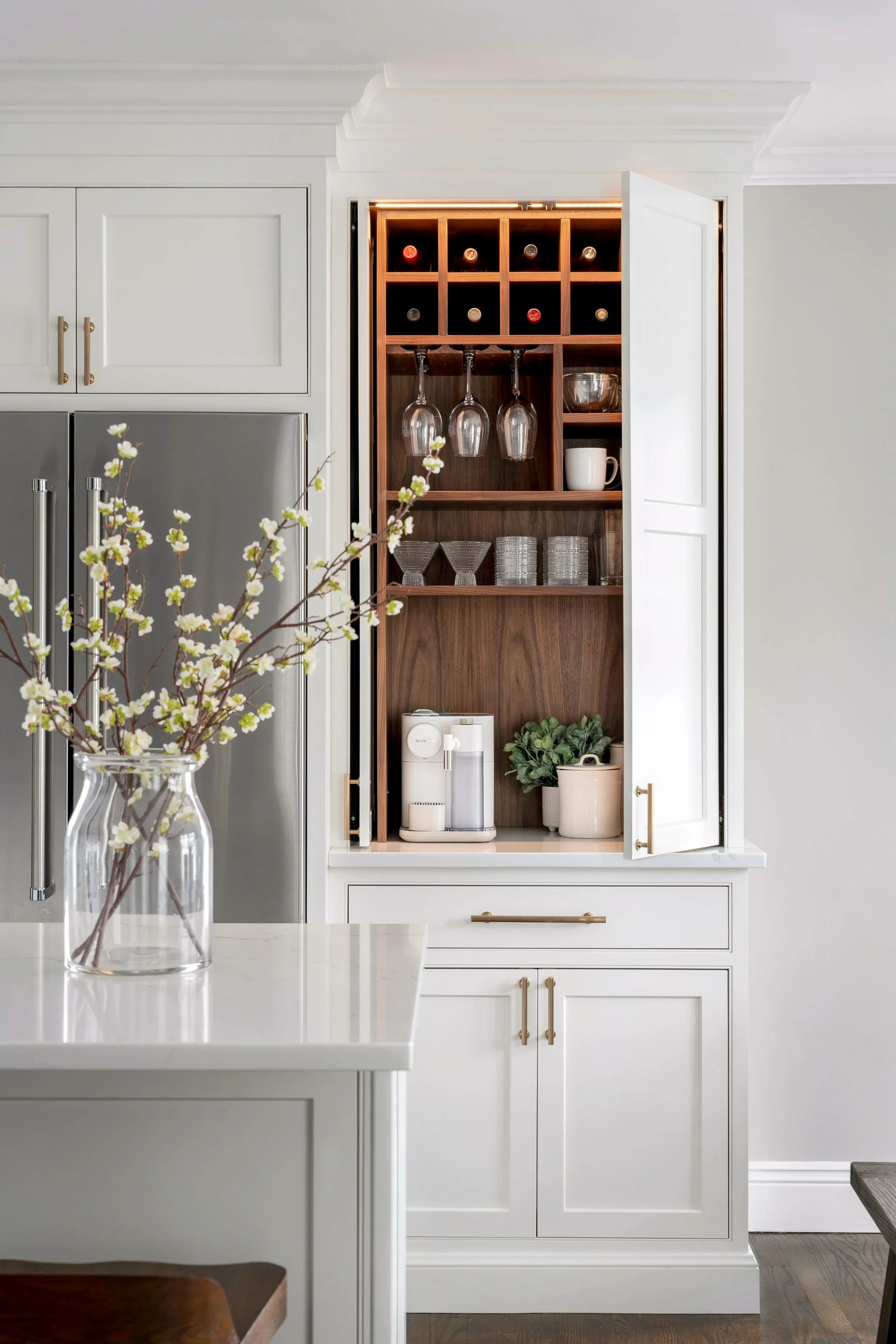 Open kitchen cabinet with wine bottles, hanging wine glasses, cups, and storage containers, next to a white countertop with a coffee machine, and a vase with white flowers on the island.