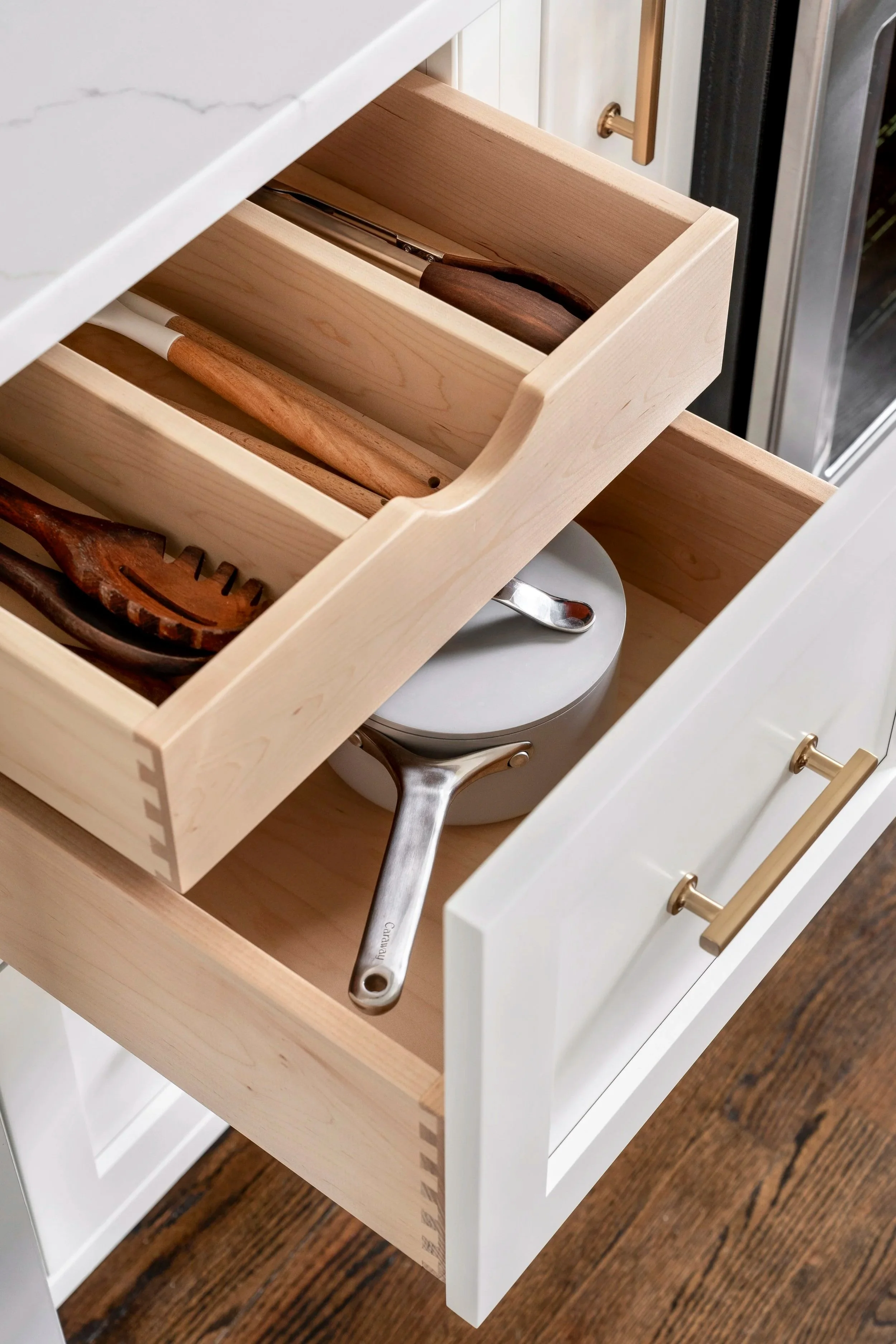 Kitchen drawer partially open containing wooden utensils and a pot with a lid and spoon, with a white cabinet and brass handles.