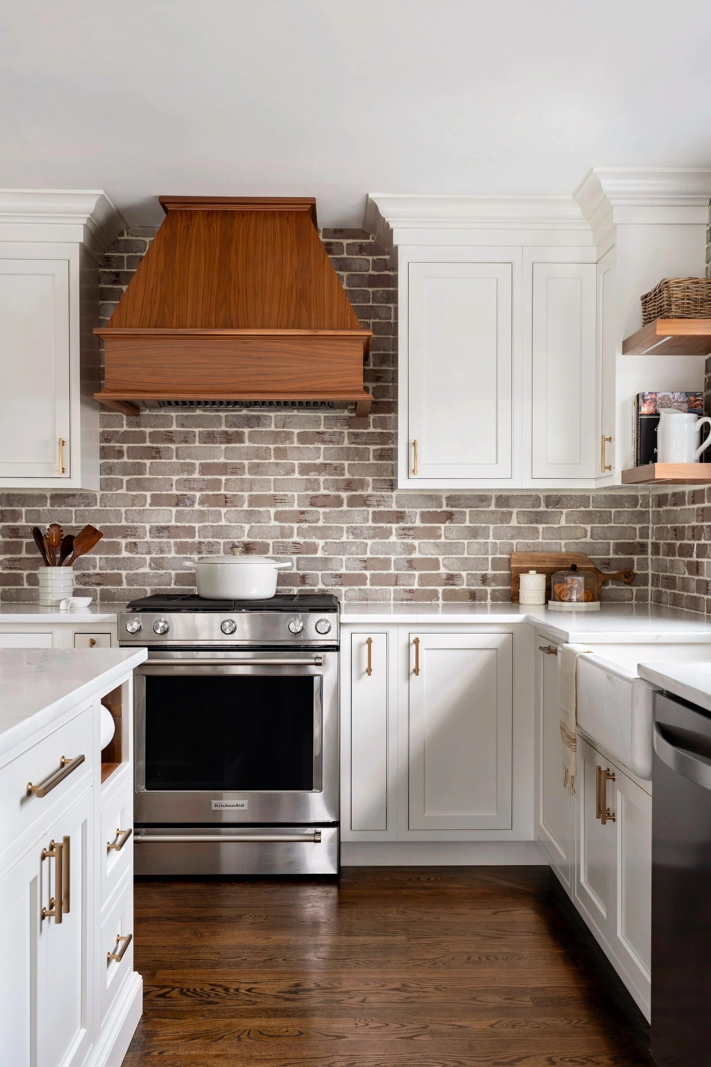 White kitchen cabinets with brass handles, stainless steel oven, brick backsplash, wooden range hood, wooden shelves, and countertop with jars and pitcher.