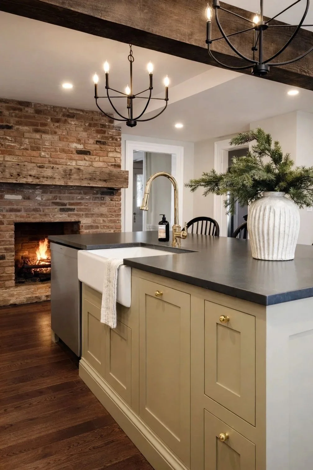 A farmhouse kitchen with soapstone countertops, island, a brass faucet, inset cabinets, sage green cabinets, wood beams, greenery, and a brick fireplace with a reclaimed wood mantle. The ceiling has recessed lighting and black chandeliers.