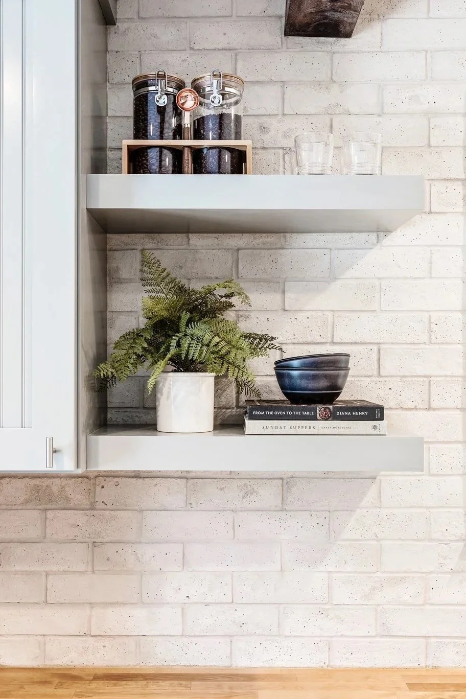 Kitchen shelf with jars of coffee beans, glasses, a potted fern, and books on a white brick wall.
