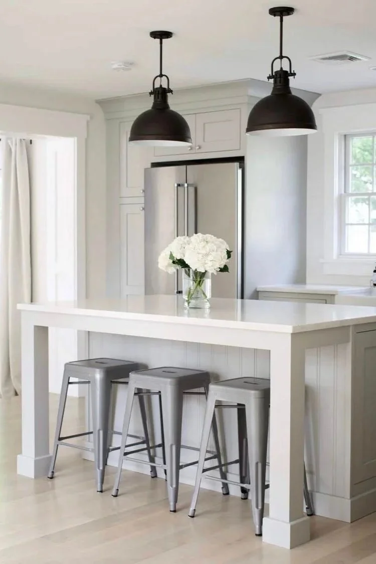 A modern kitchen with a white island, three silver metal bar stools, black pendant lights, white cabinetry, a stainless steel refrigerator, a window with white curtains, and a glass vase with white flowers on the island.