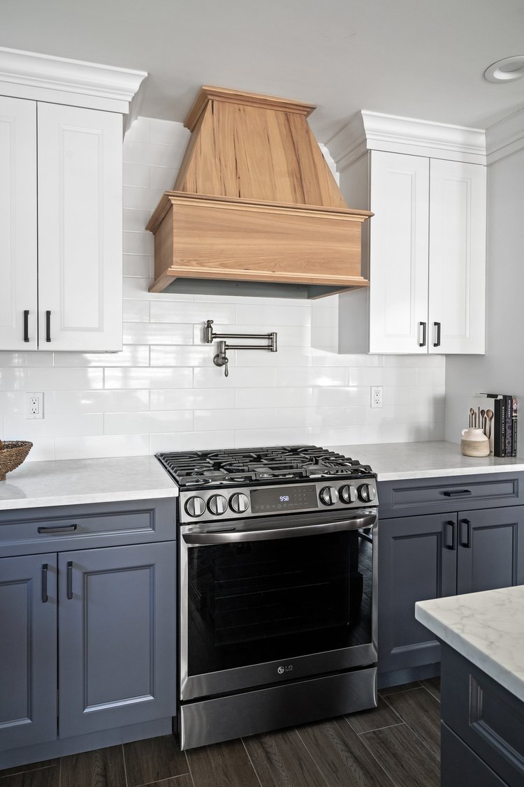 A modern kitchen with white upper cabinets, gray lower cabinets, a stainless steel oven, a white marble countertop, a wooden range hood, and white subway tile backsplash.