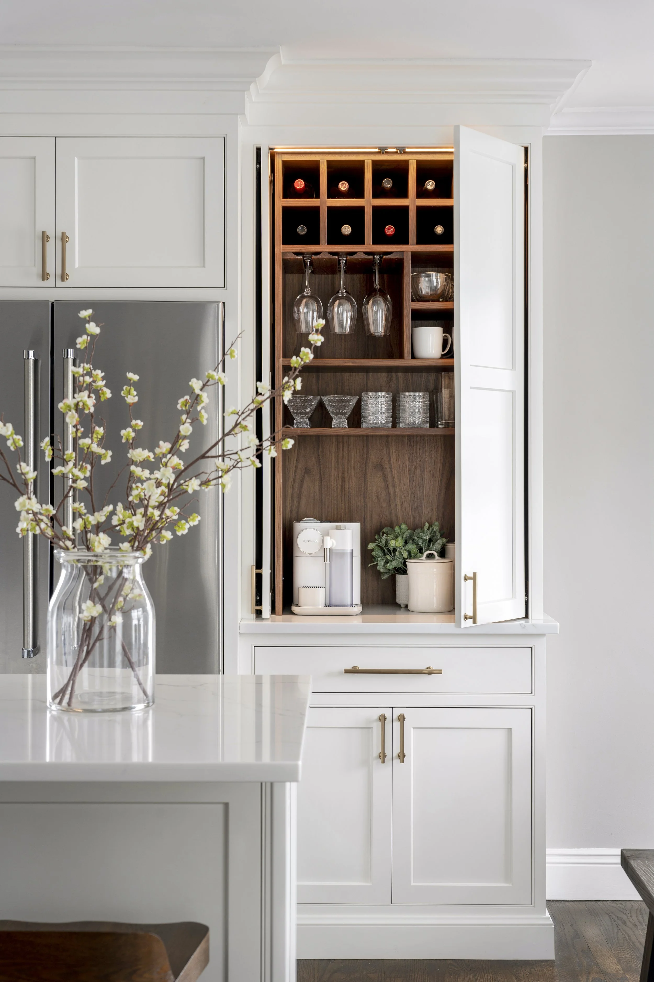 Open kitchen cabinet with wine bottles, hanging wine glasses, cups, and storage containers, next to a white countertop with a coffee machine, and a vase with white flowers on the island.
