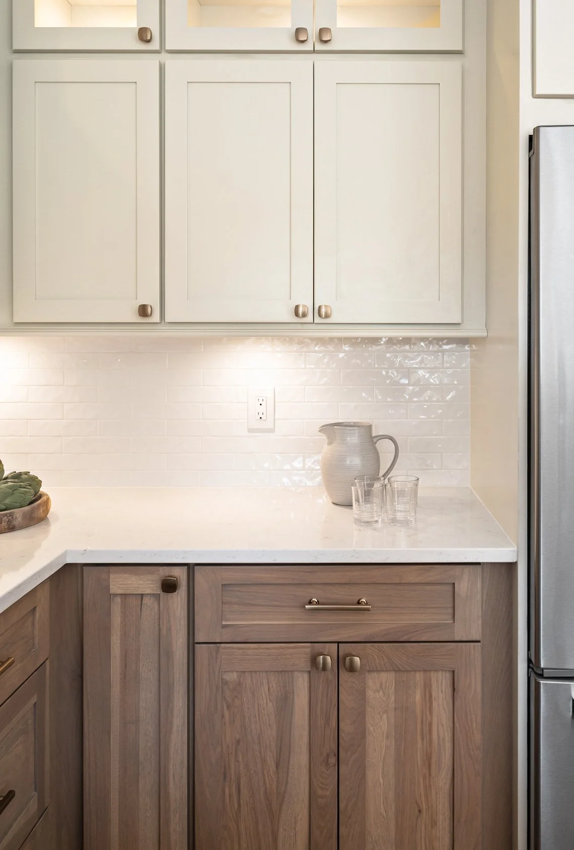 Kitchen counter with white upper cabinets, light-colored backsplash, beige pitcher, and two clear glasses, with a stainless steel refrigerator on the right.