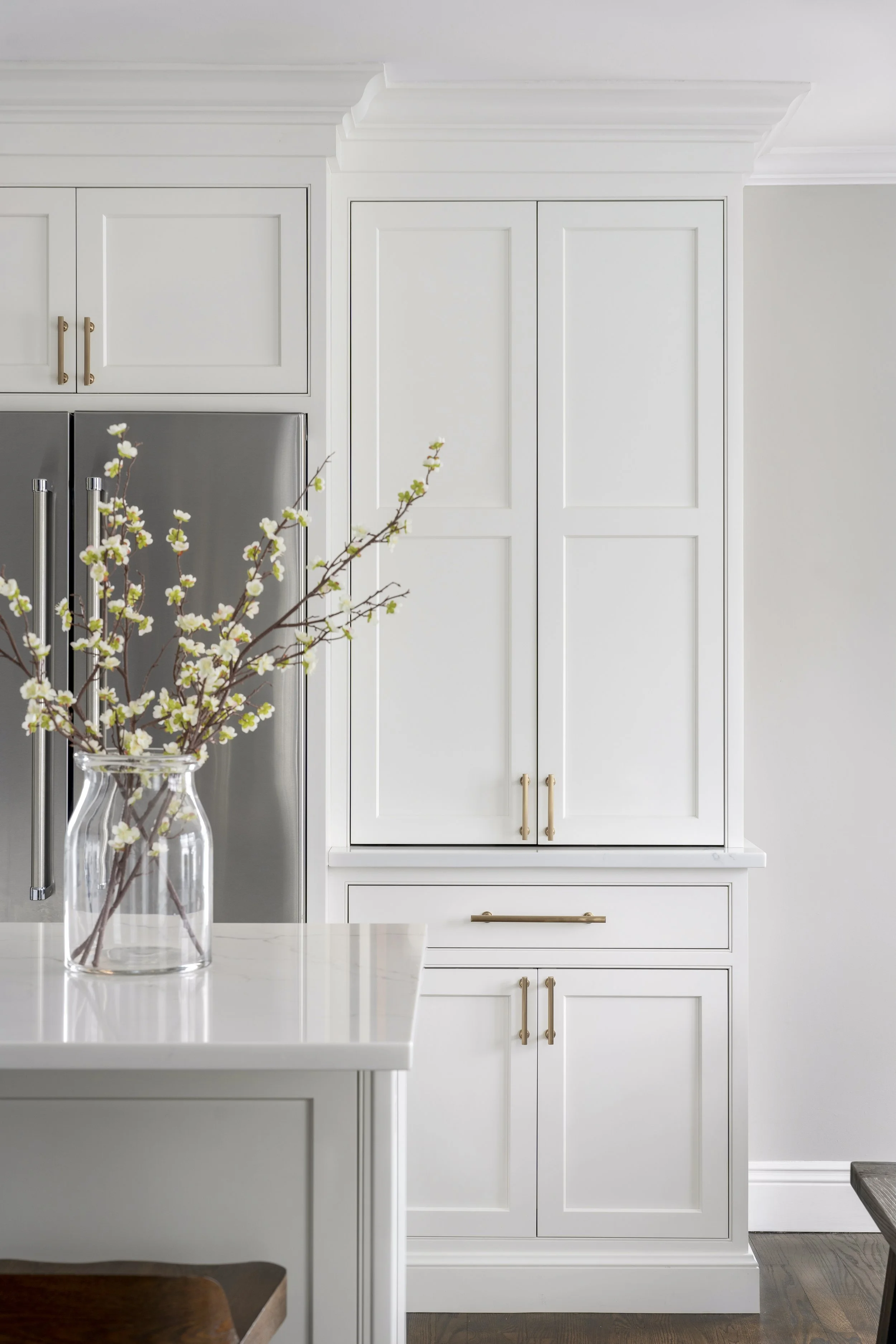 A white kitchen with white cabinets, a white countertop, and a decorative crown molding. A glass vase with white flowering branches is on the counter. There is a stainless steel refrigerator with vertical handles on the left side of the image. The floor is wood.
