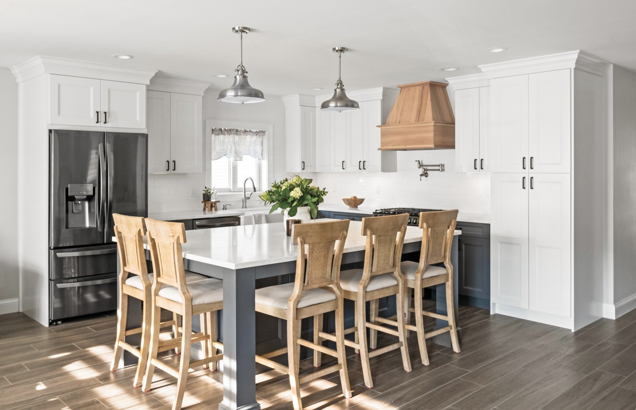 Modern kitchen with white cabinets, a dark island, stainless steel refrigerator, and a wooden range hood. There are five wooden bar stools around the island, and a flower arrangement on top. The kitchen has wood flooring and pendant lights.